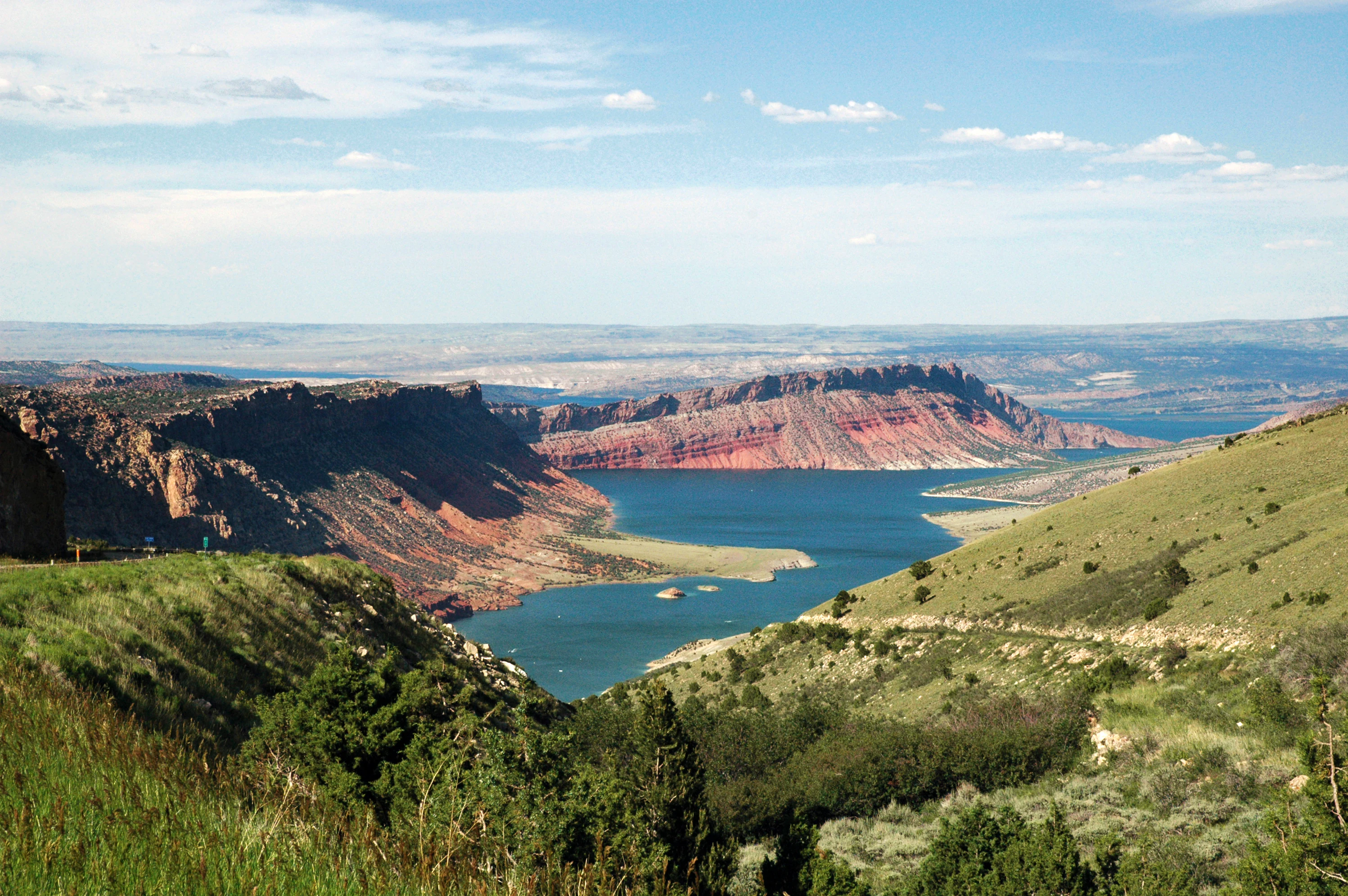 A high vantage view of a blue reservoir nestled between high red sandstone cliffs and sloping green, shrub-covered hillsides under a partly cloudy sky.