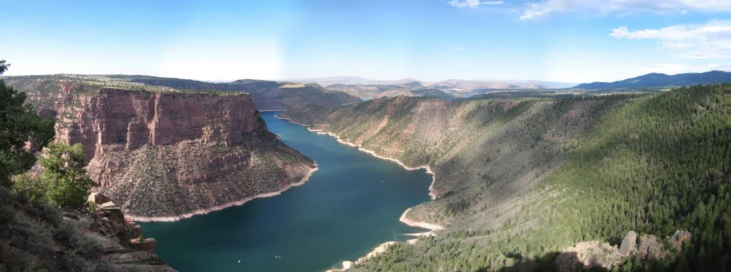 A wide-angle landscape view showing a large reservoir winding through a canyon with steep, reddish-brown cliffs on one side and a forested slope on the other.