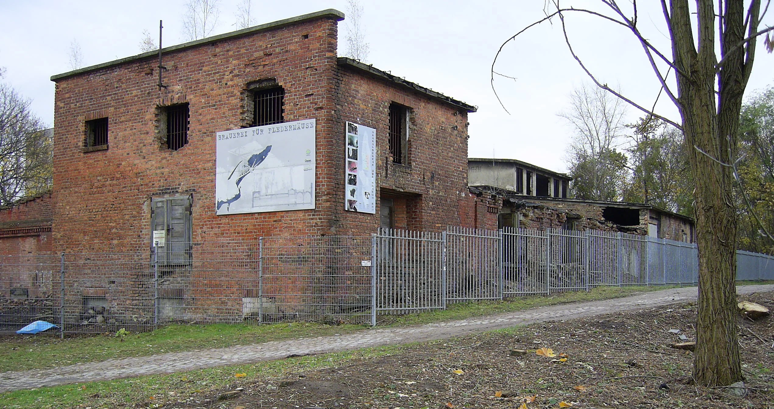 A weathered red brick industrial building with a fenced perimeter and informative signs mounted on the wall.