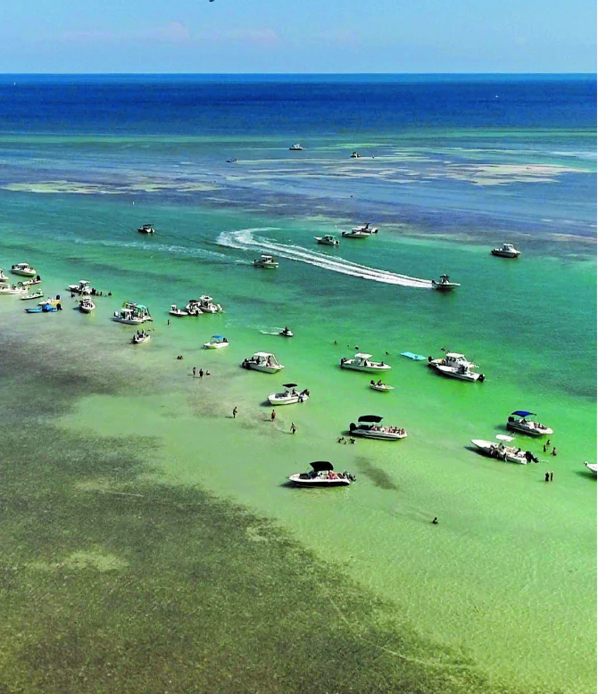 An aerial view of many small motorboats and pontoons gathered in very shallow, clear, turquoise water, with people wading nearby.