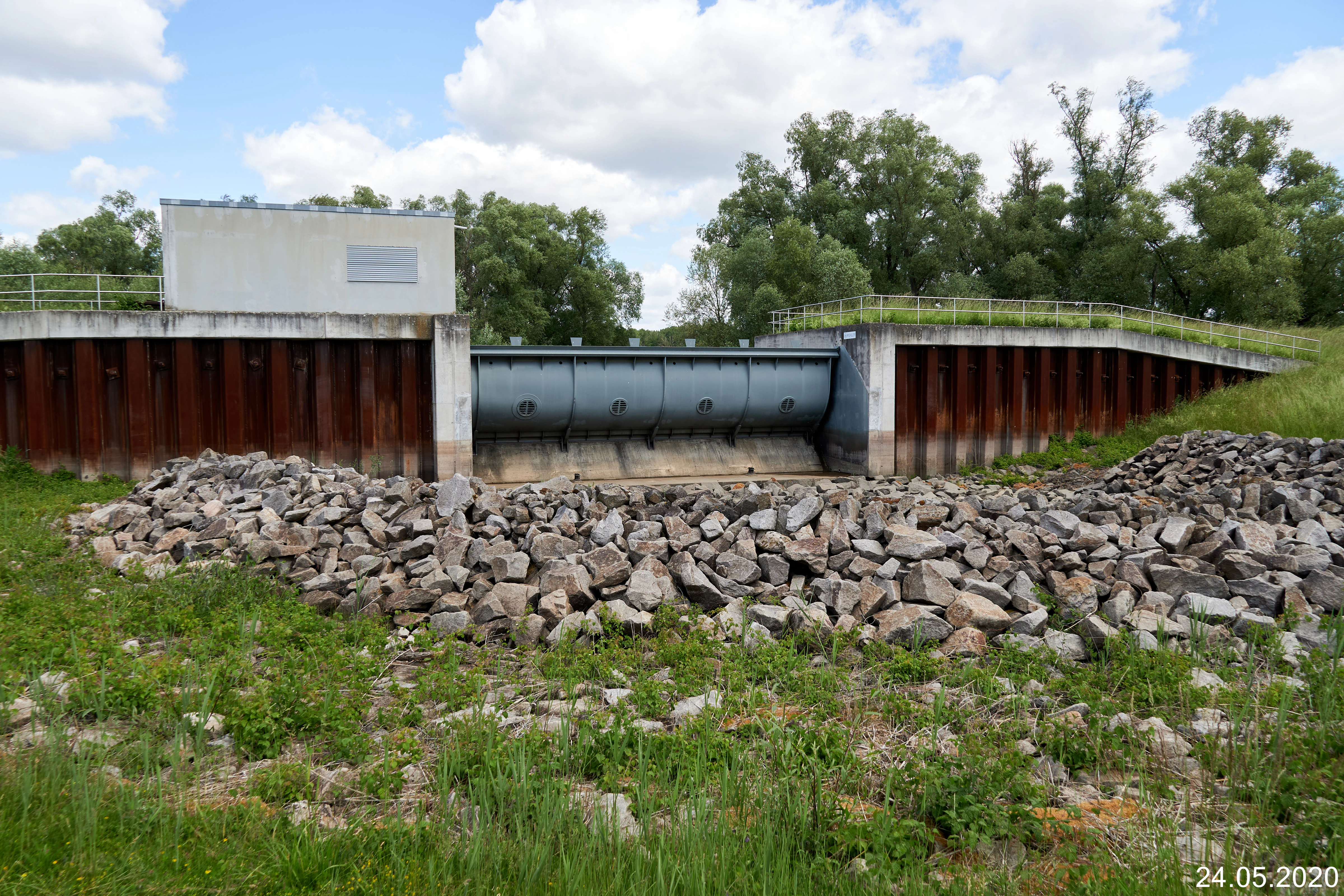 A wide eye level shot of the concrete and steel north gate structure of the Flotzgrün polder with a pile of grey rocks in the foreground and trees in the background.