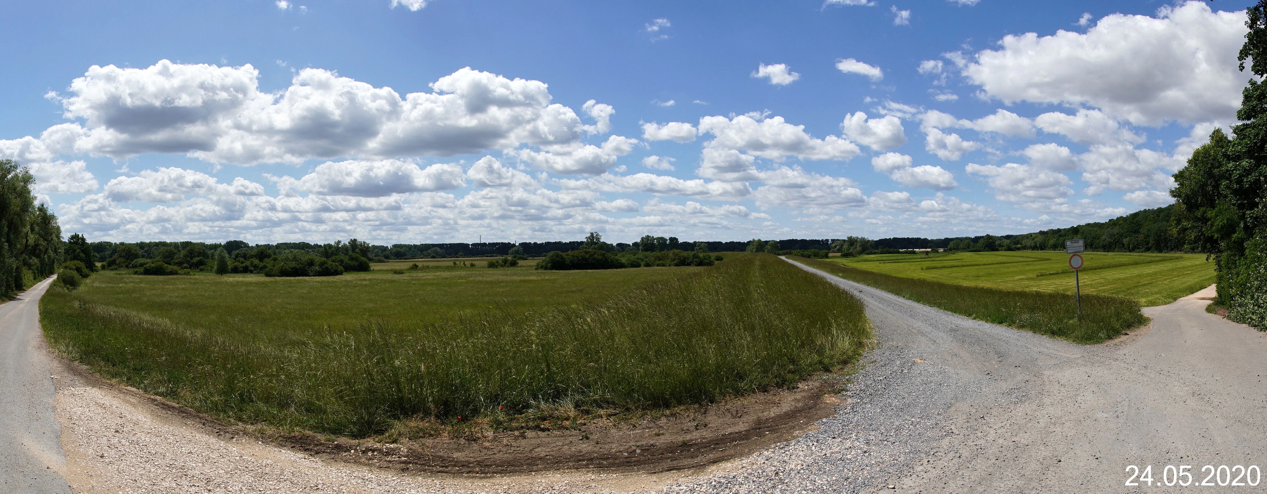 A wide panoramic landscape shot of Flotzgrün showing grassy fields, gravel paths, and a blue sky with scattered clouds.