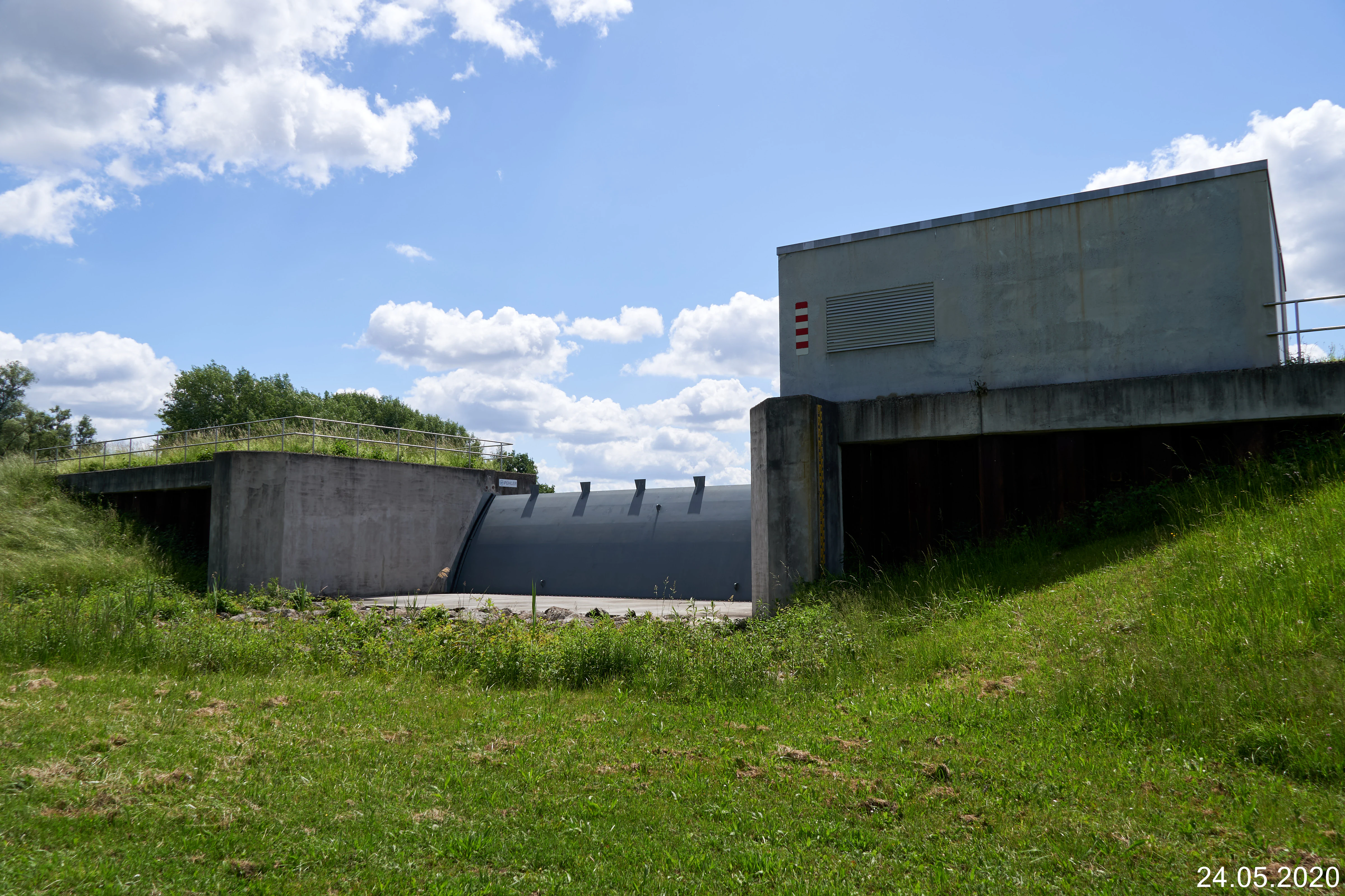 A concrete flood control building and sluice gate structure set into a grassy hillside on a clear day.