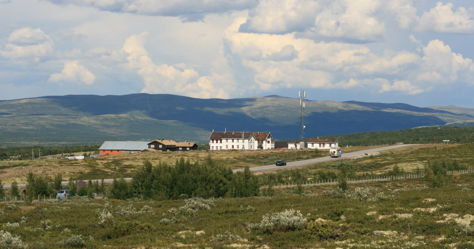 A wide view of the Fokstugu mountain lodge complex, characterized by several traditional buildings set within a rolling, shrub-covered moorland with mountains in the background.