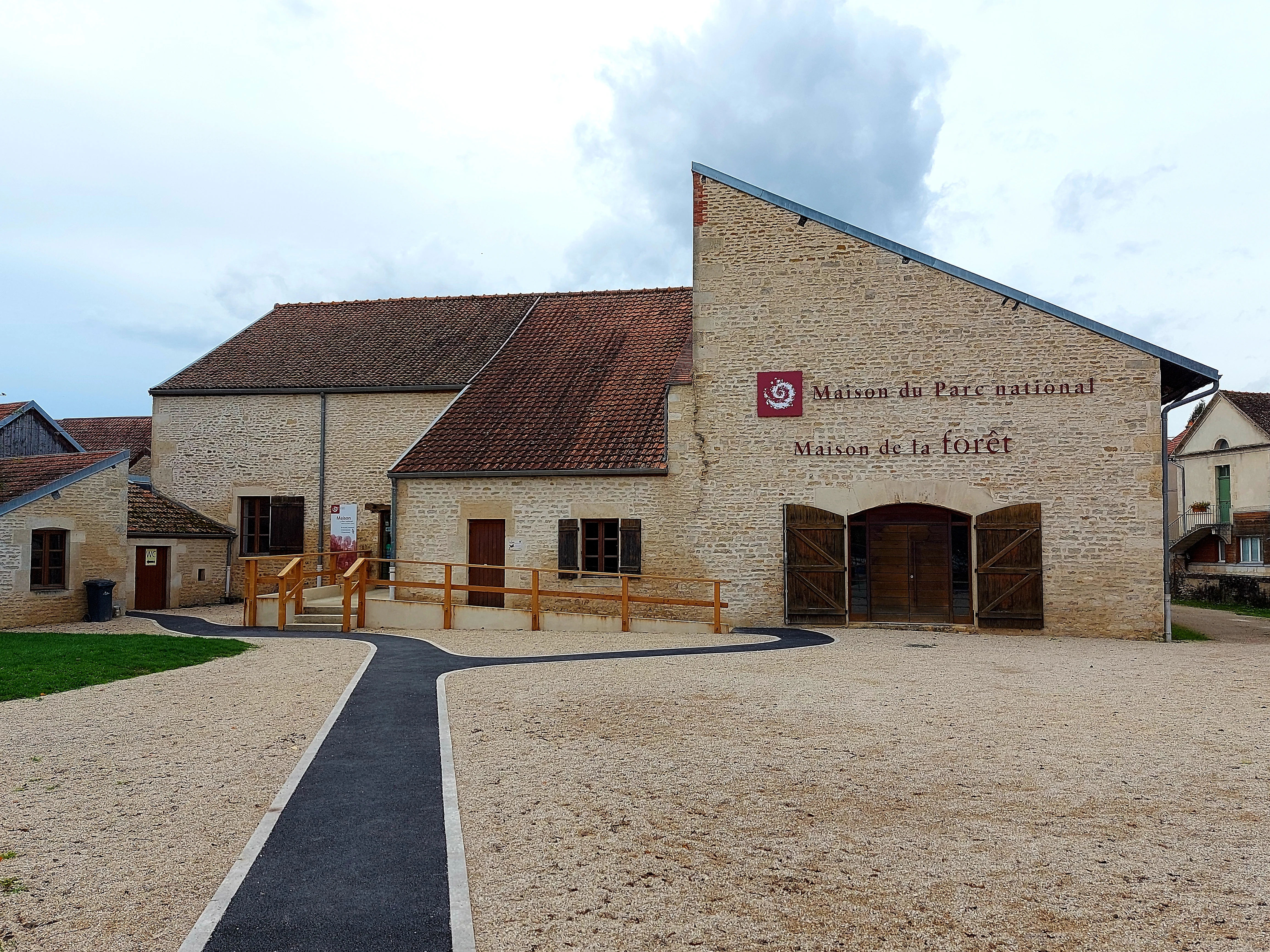 Stone building with brown roof and red circular emblem, labeled 'Maison du Parc national de forêts' and 'Maison de la Forêt,' with gravel courtyard and paved pathway