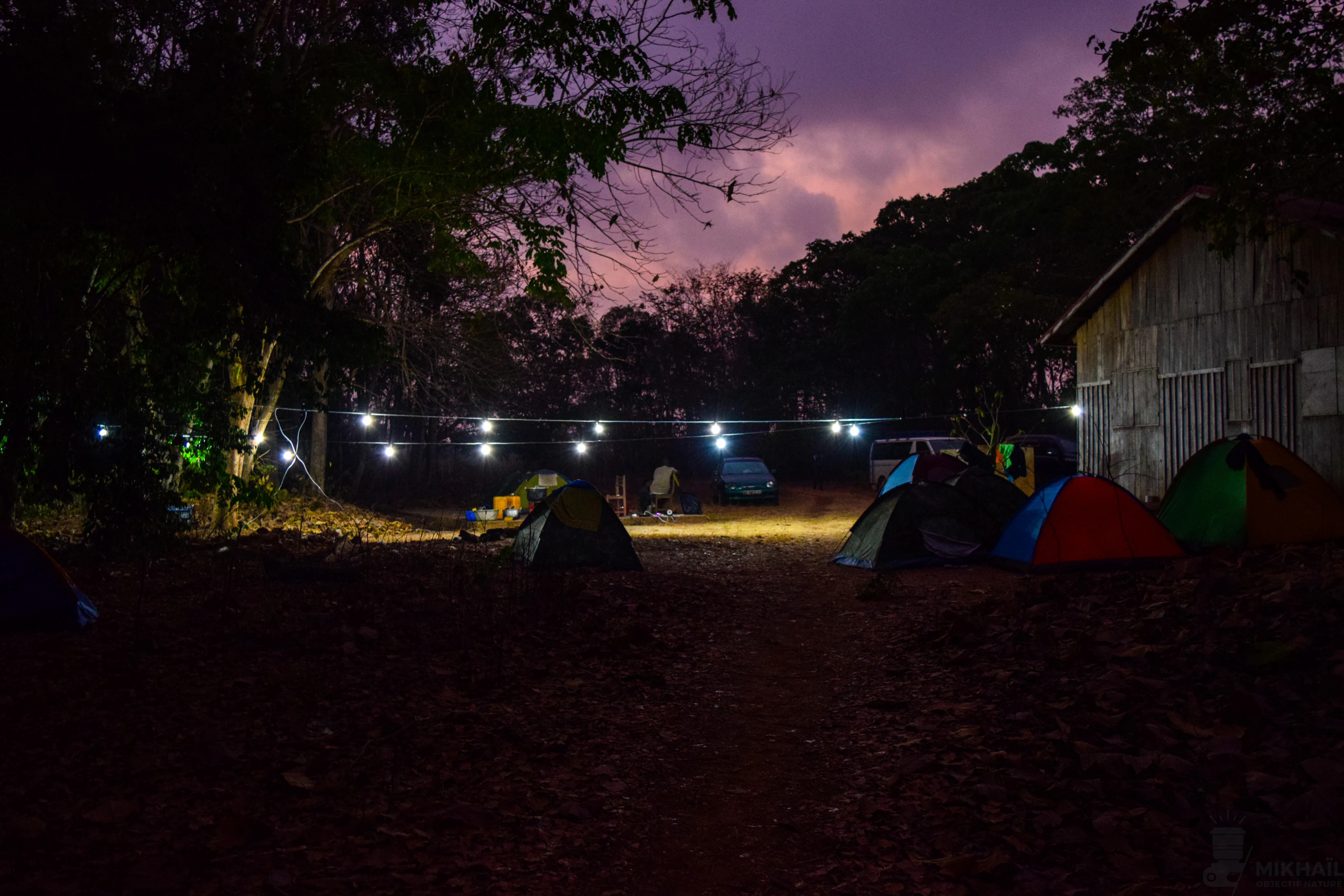 A base camp area with multiple colorful camping tents set up on dirt ground at dusk, illuminated by a string of outdoor lights, with a wooden building visible on the right.
