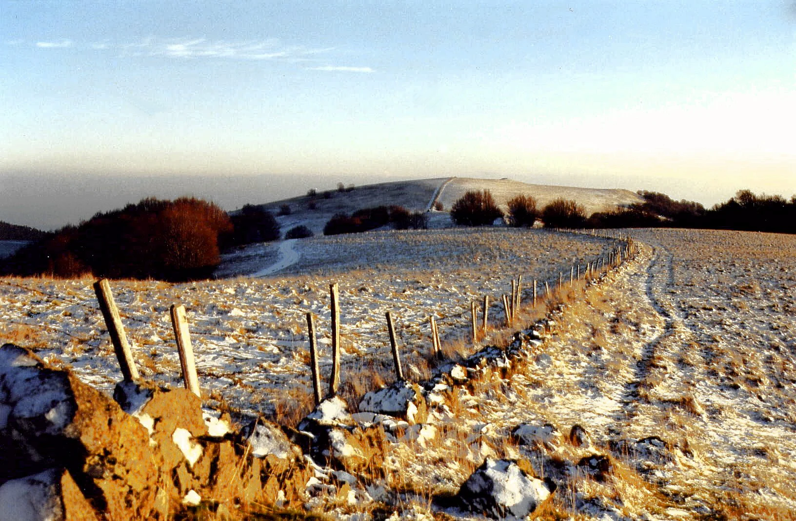 A wide view of a snowy hillside landscape with a stone wall and wooden fence stretching into the distance towards a rolling summit under a pale sky.