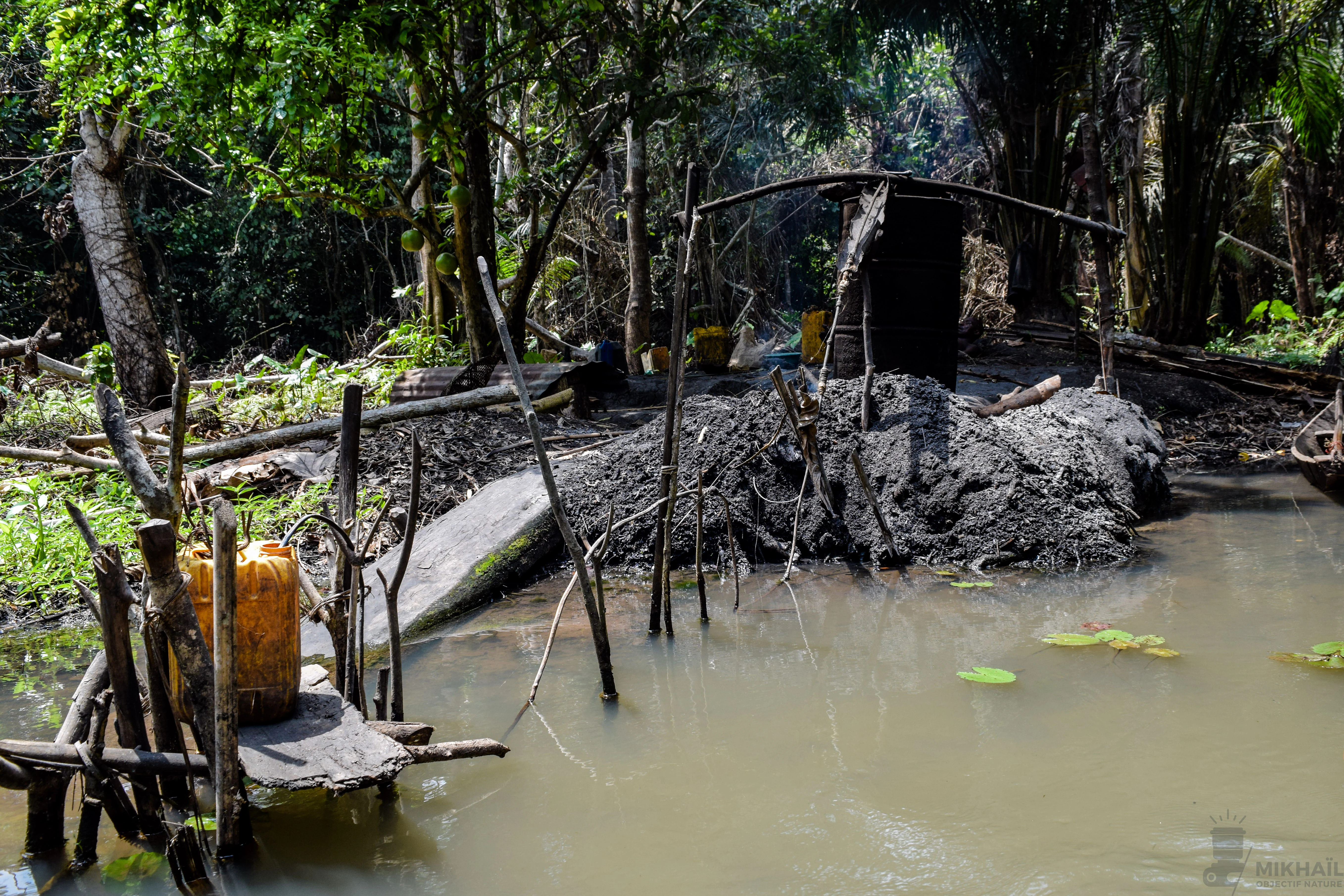 A small, rustic outdoor processing station for raphia alcohol production, featuring a muddy mound, wooden structures, and yellow plastic containers, situated along a dark, shallow waterway in a dense forest.