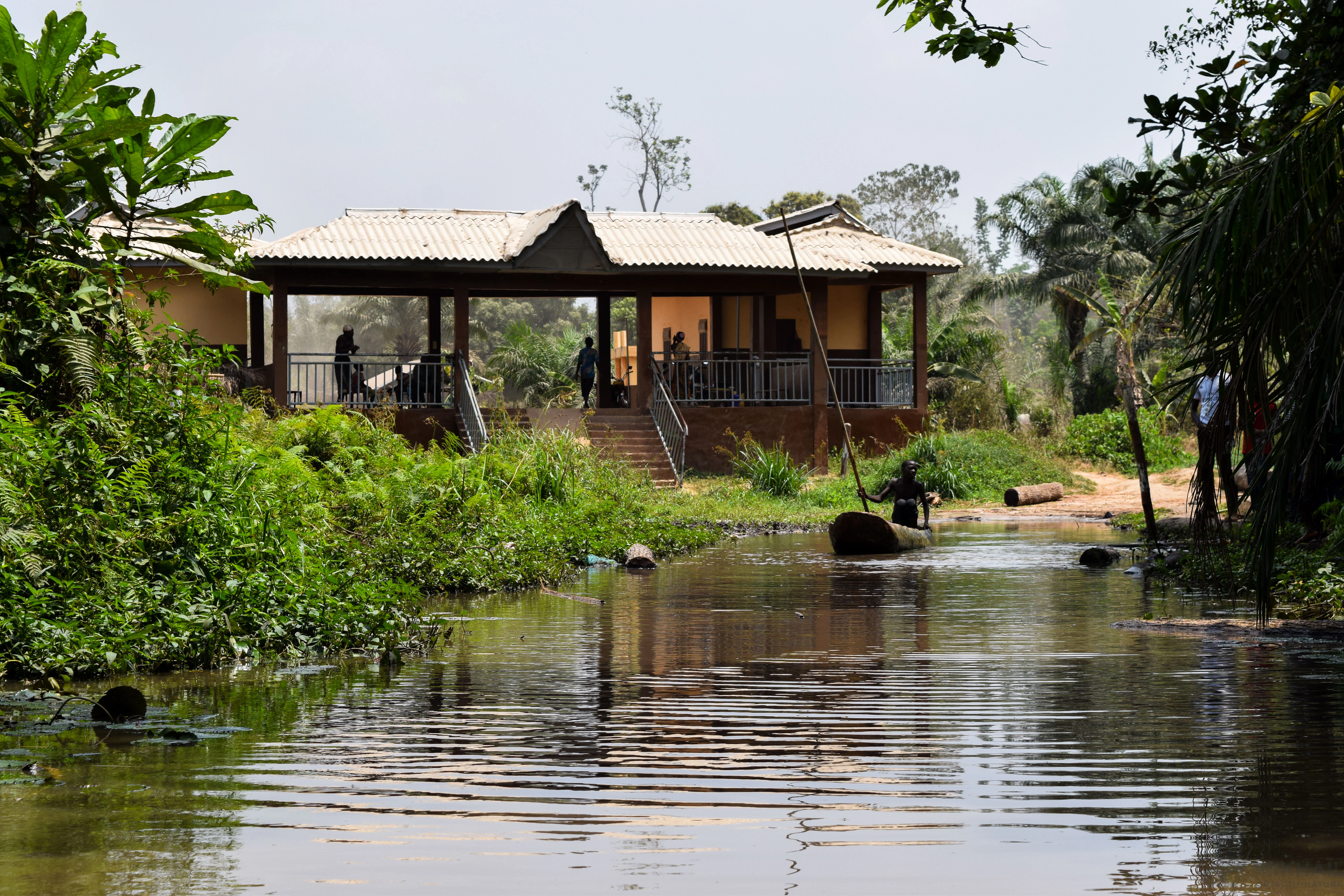 A small building with a covered porch and stairs overlooks a narrow, dark waterway in a forest, with a person in a wooden dugout canoe nearby.