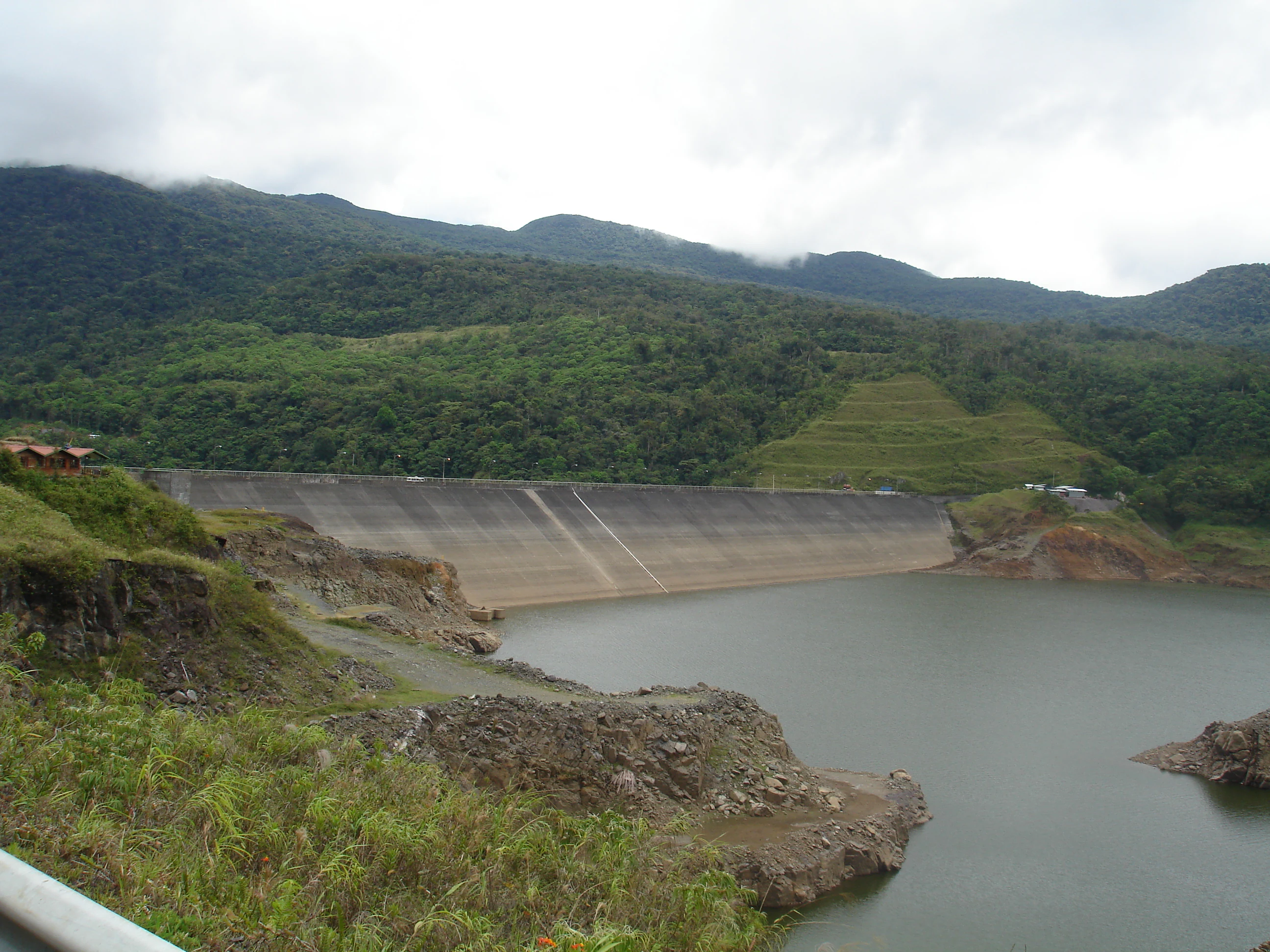 A wide shot of a large concrete dam wall holding back a reservoir of water, surrounded by lush green hills and a forested mountain range under a cloudy sky.