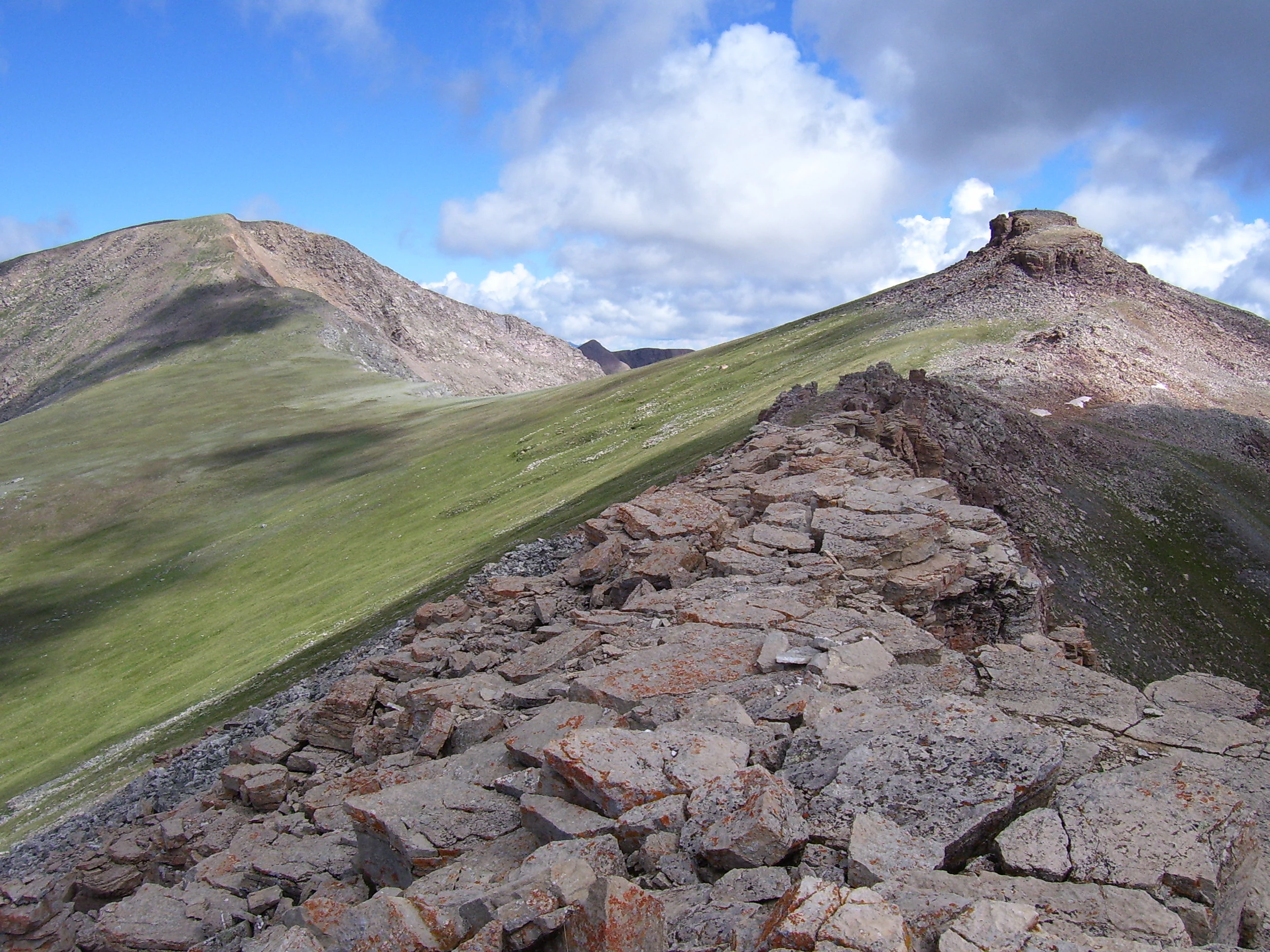 A view along a rocky ridge with loose stones in the foreground, ascending a green slope towards two mountain peaks under a partly cloudy sky.
