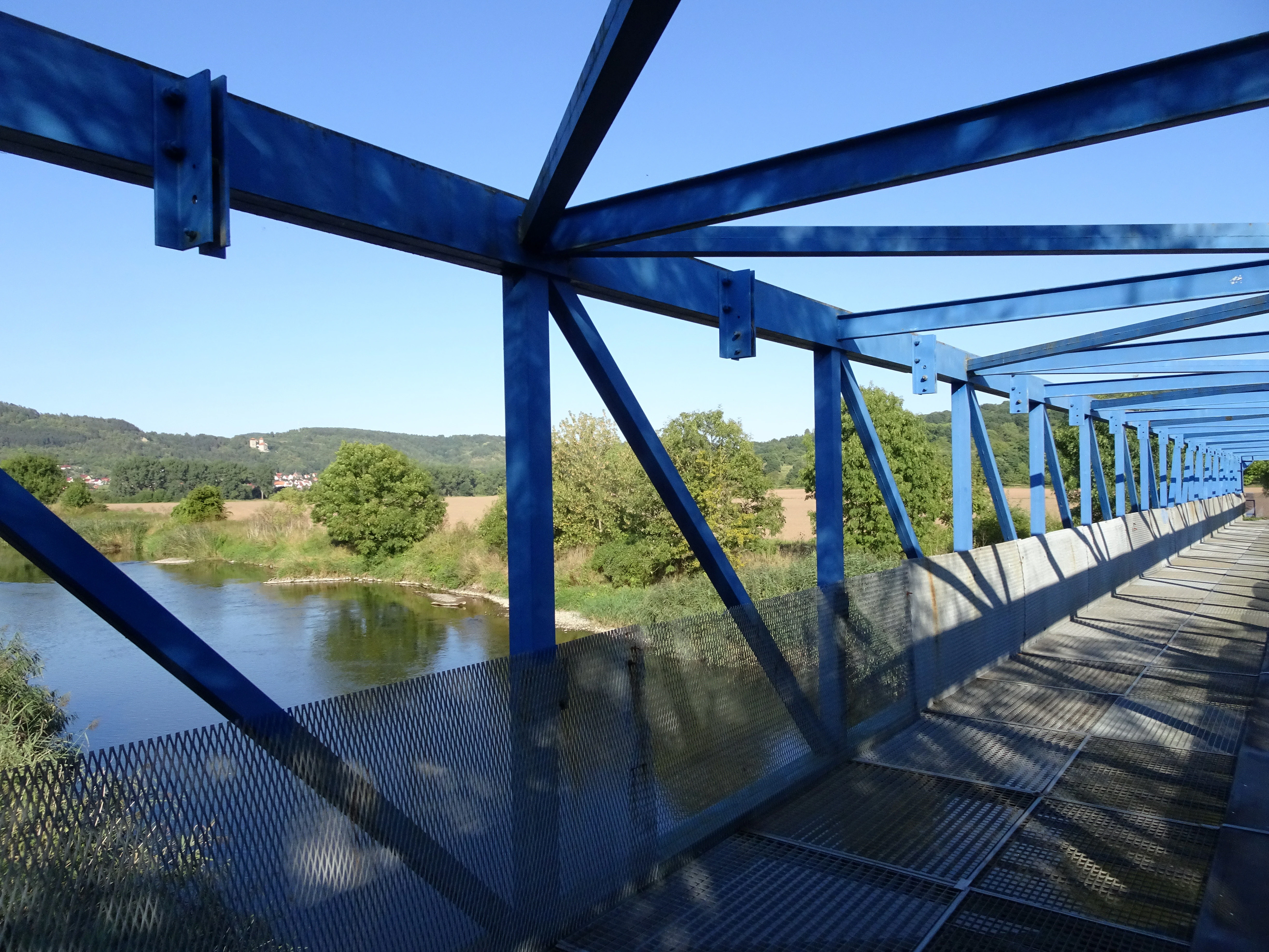 A blue metal truss pedestrian bridge with mesh walkway extending into the distance, spanning a river with green trees and rolling hills visible in the background under a clear blue sky.