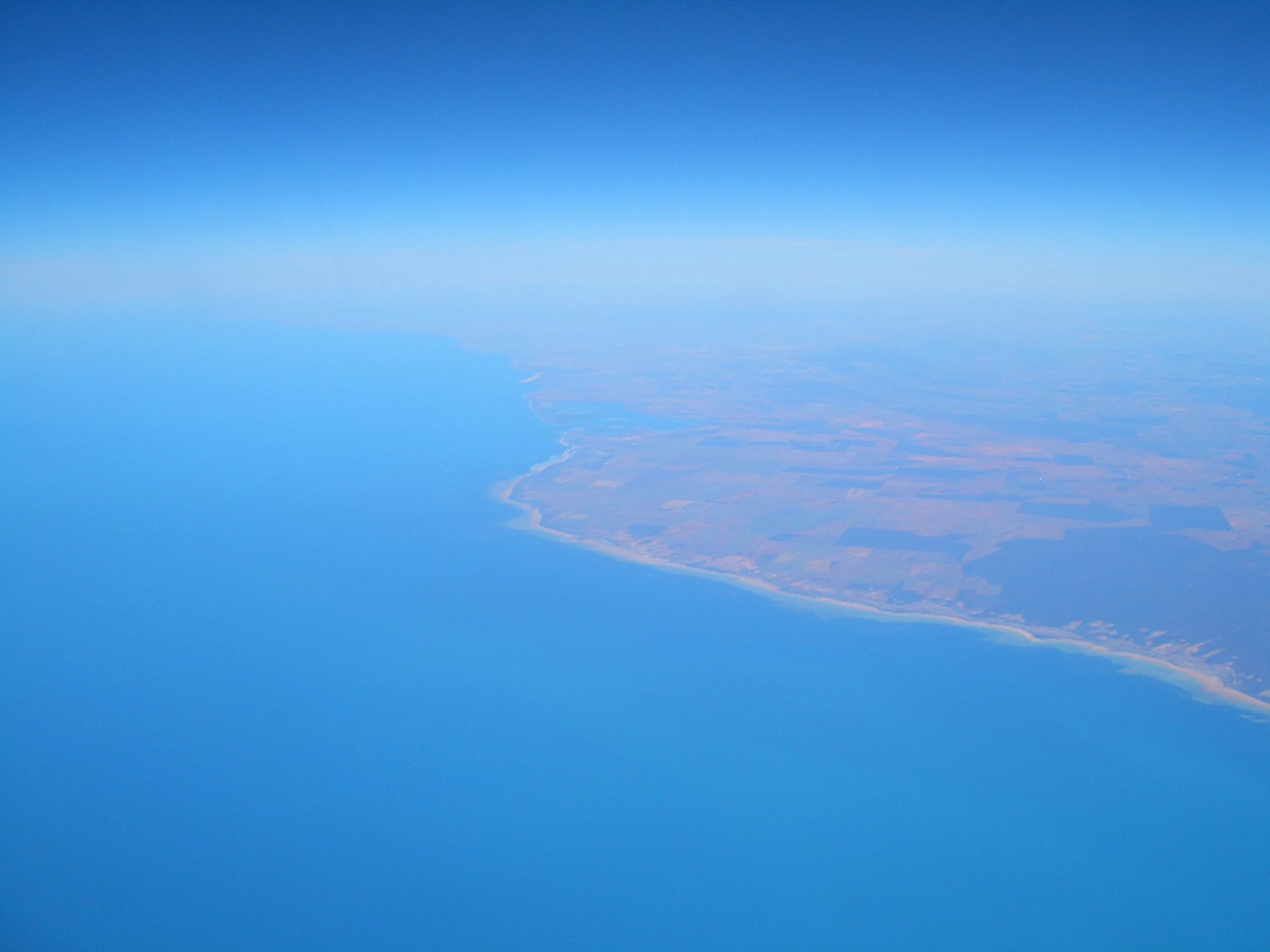 High altitude aerial view of the coastline where blue ocean water meets a tan and brown coastal landscape.