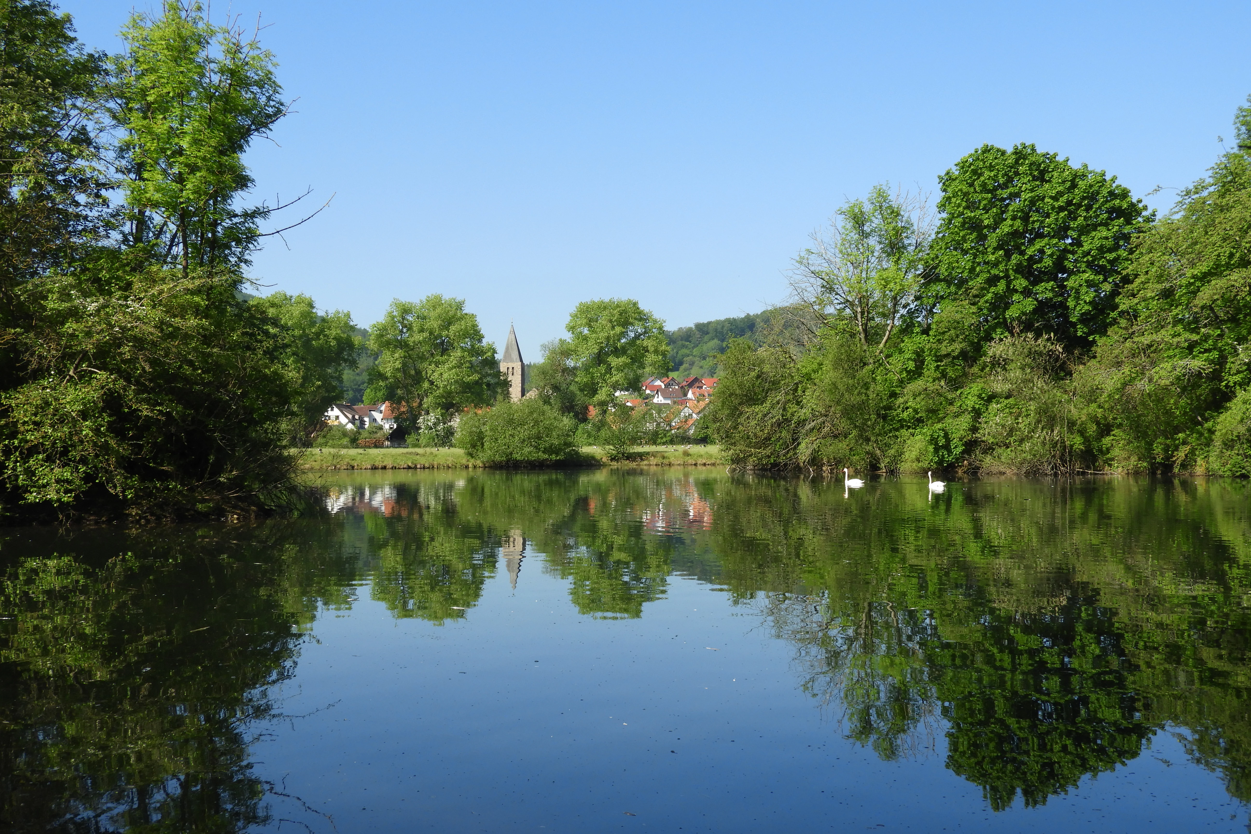 A calm lake reflects lush green trees and a clear blue sky, with the steeple of a distant village church visible in the background.