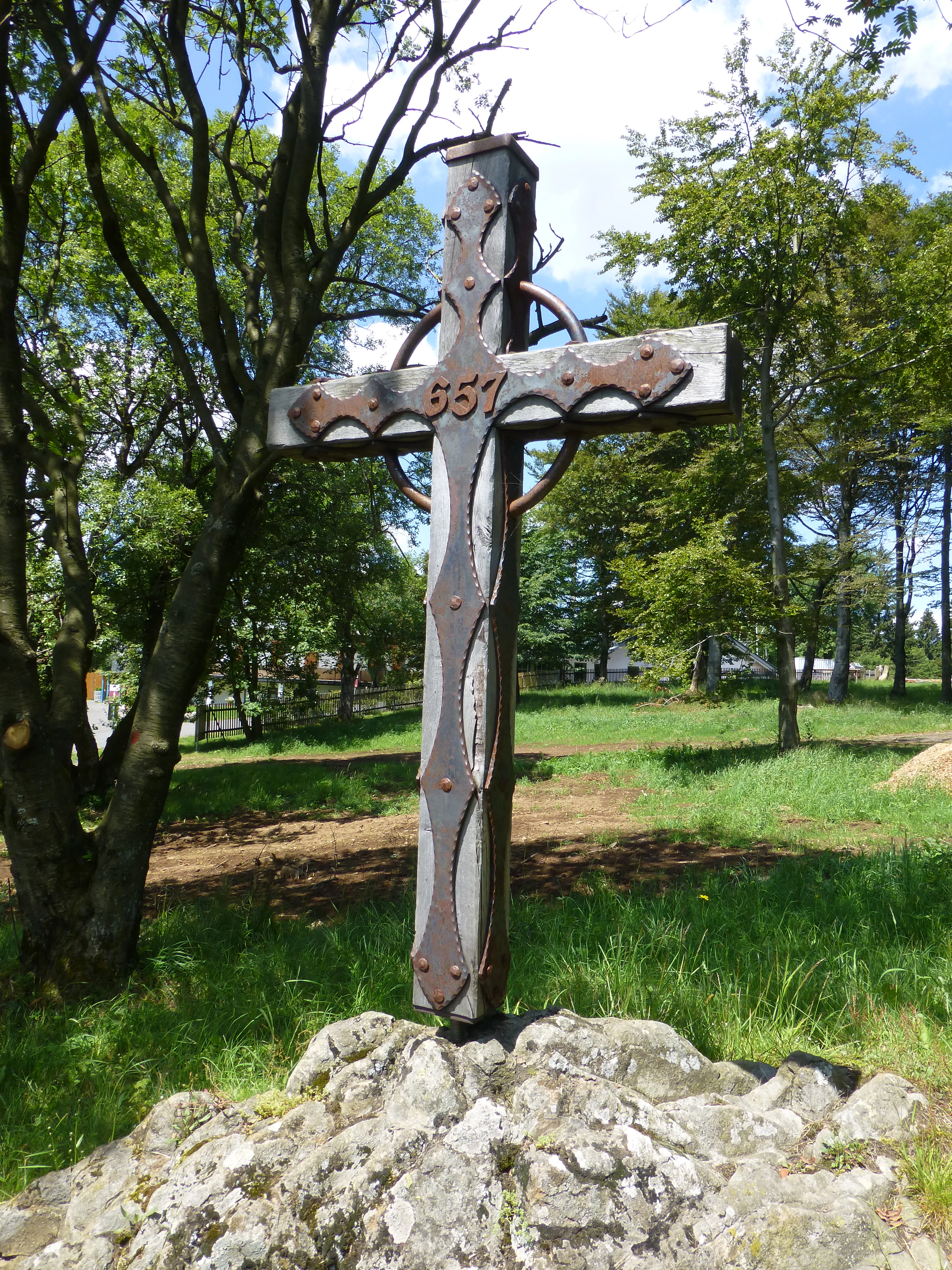 A weathered wooden summit cross decorated with metal plating and the number 657, mounted on a rocky basalt base in a grassy, forested area.