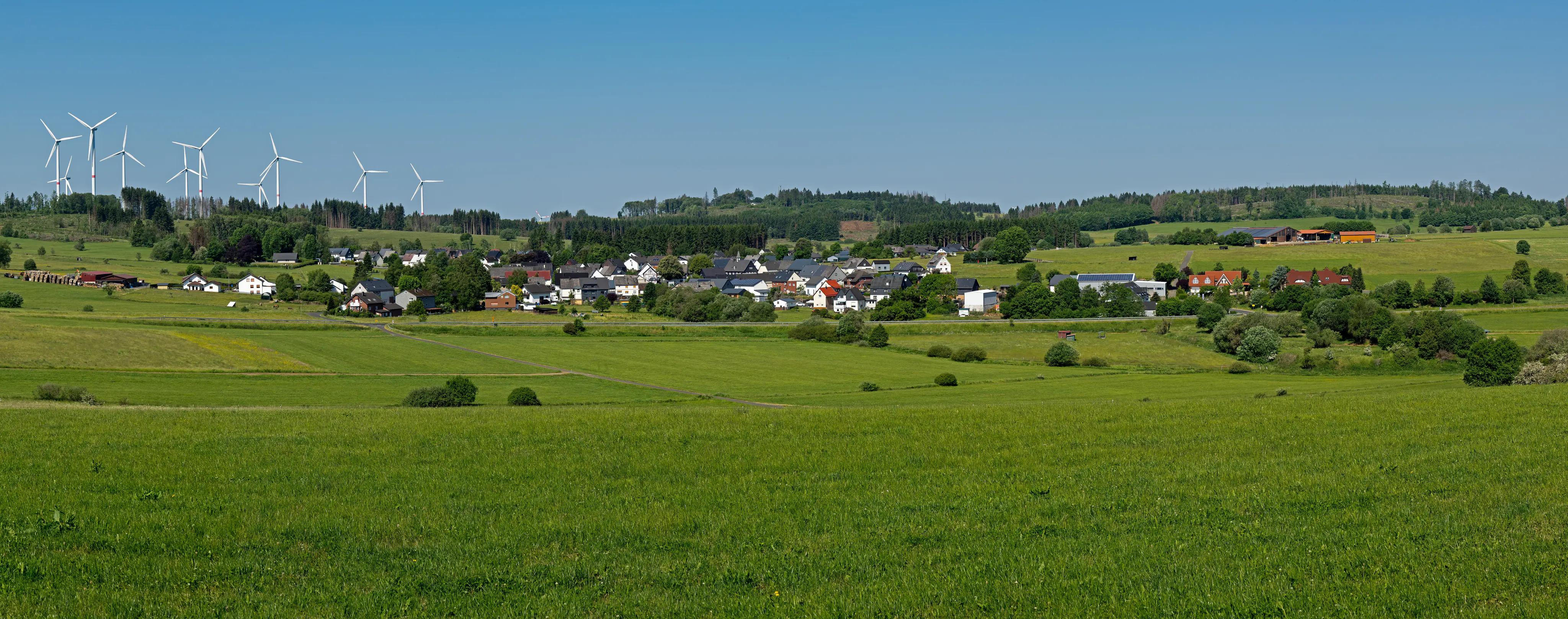 Panoramic landscape view of a rural village nestled in rolling green hills with wind turbines visible in the distance under a clear blue sky.