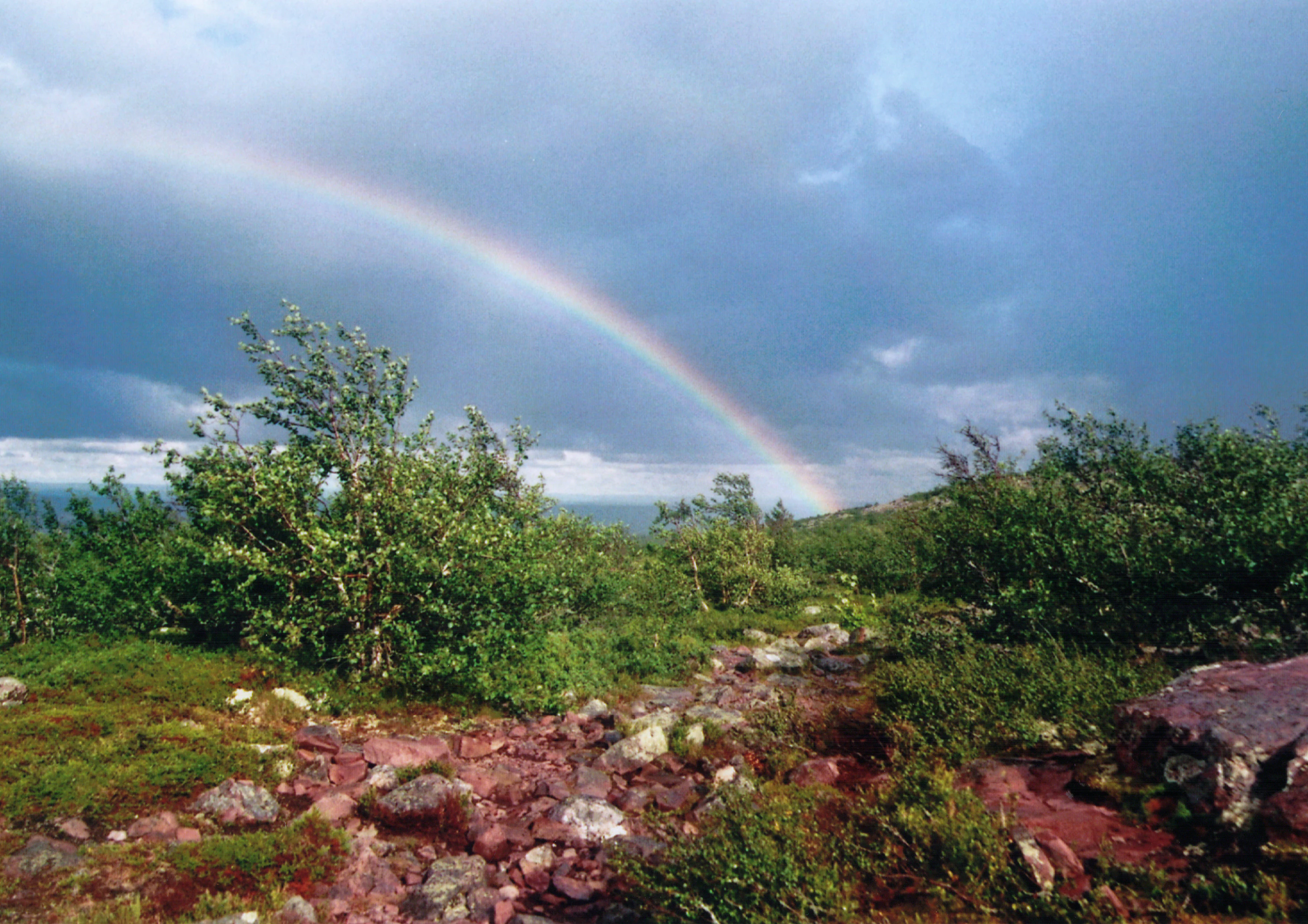 Landscape with rainbow over rocky terrain, green vegetation, and cloudy sky