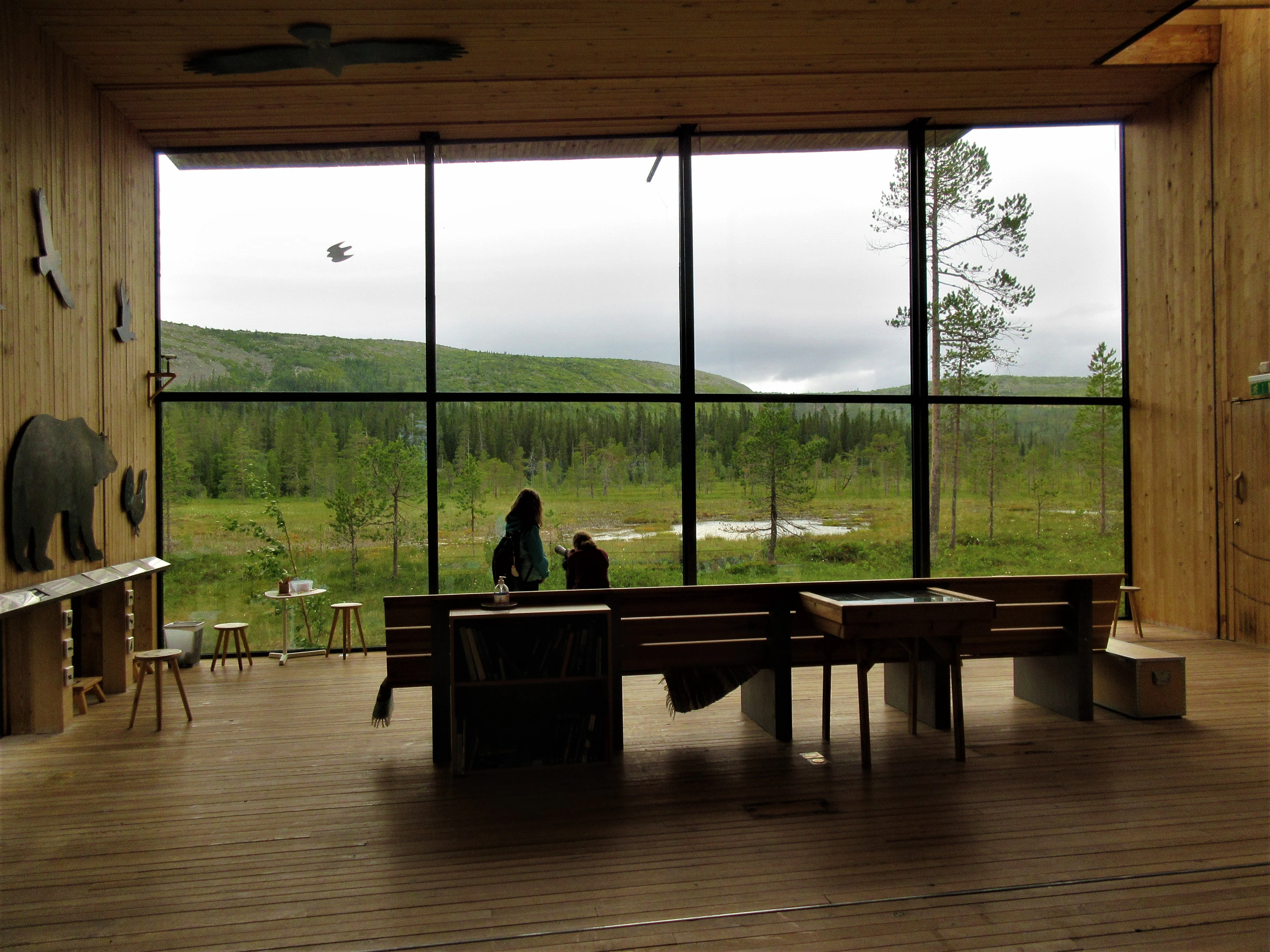 Wooden interior of Naturum Fulufjället visitor center showing large windows with a view of grassy fields and mountains outside. Two people are seated in the foreground with silhouettes visible against the bright exterior.