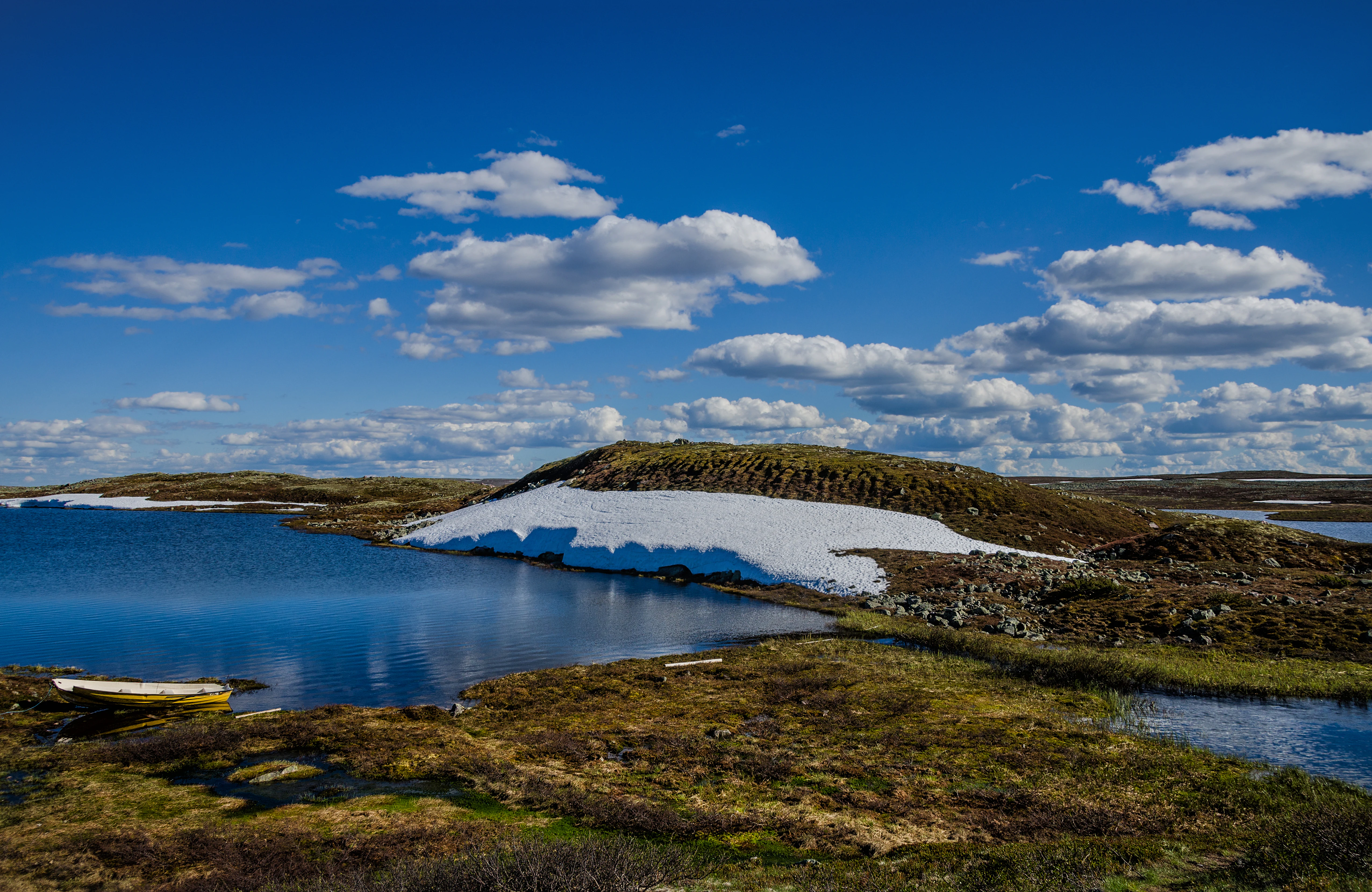 Snowmelt lake with grassy terrain, snow patches on a hill, and a yellow boat on the shore under a blue sky with scattered clouds