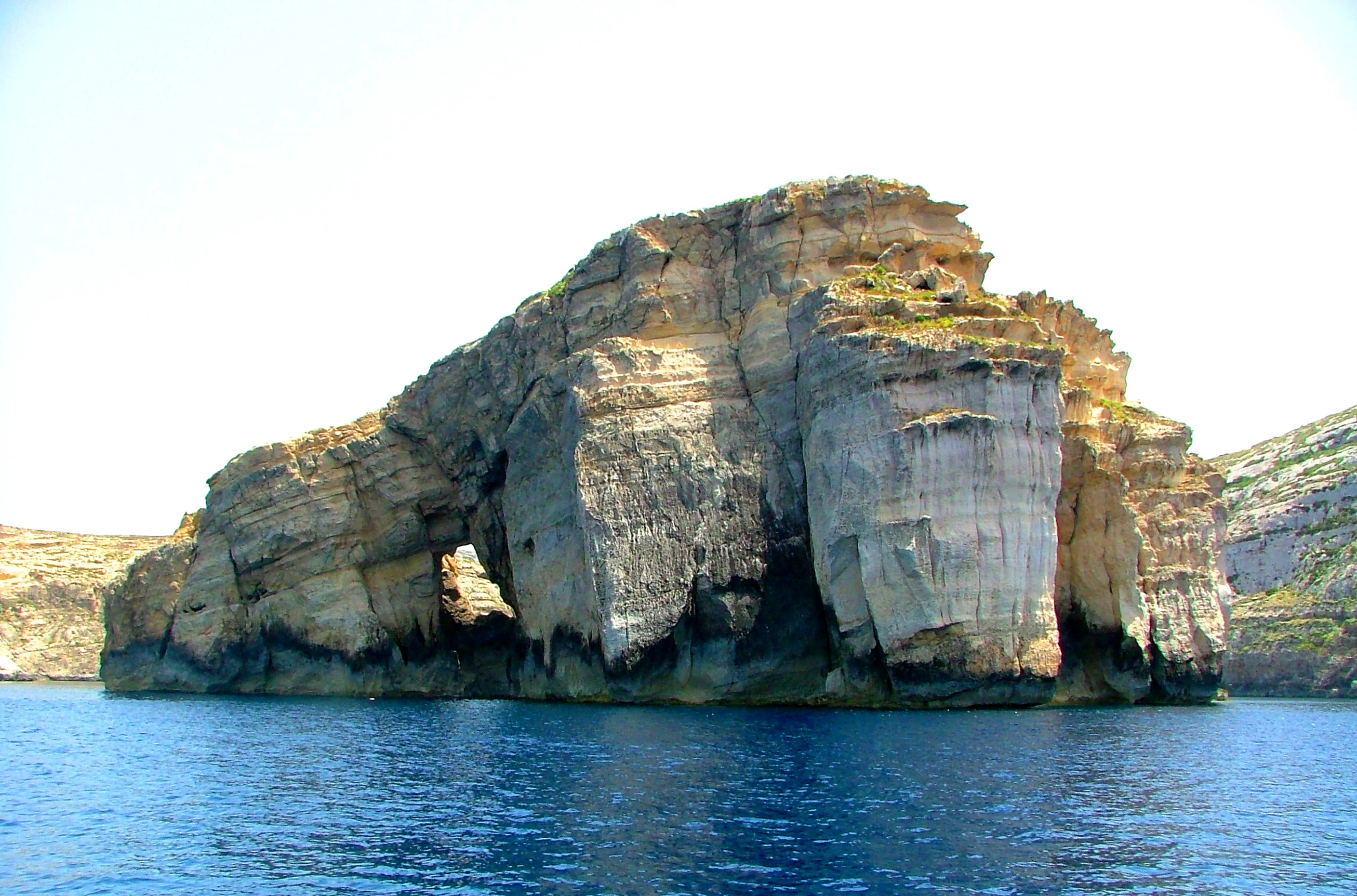 A large, weathered limestone islet with a natural archway, rising from deep blue water under a bright sky.