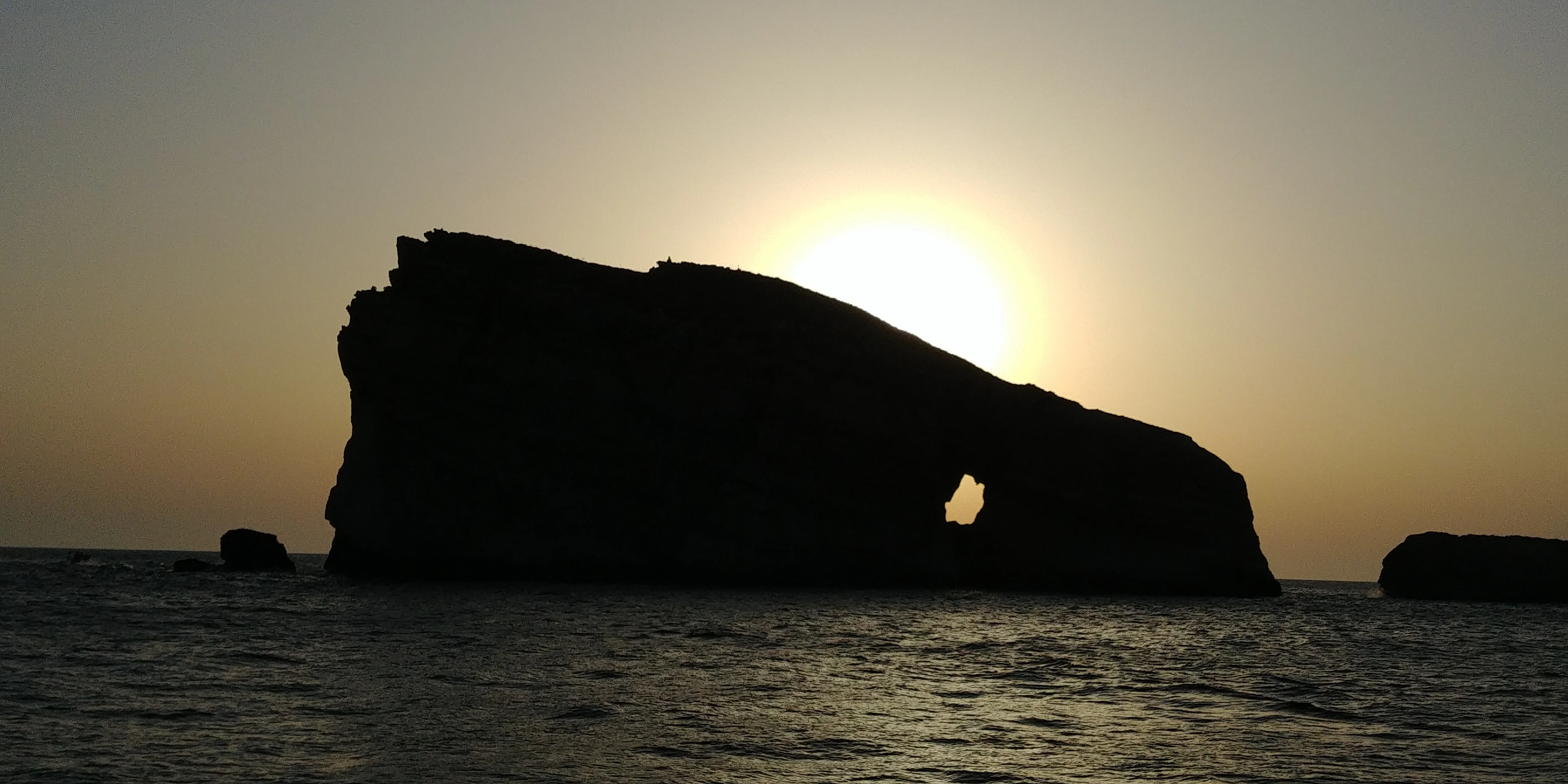A backlit silhouette of Fungus Rock limestone islet surrounded by the sea during sunset.