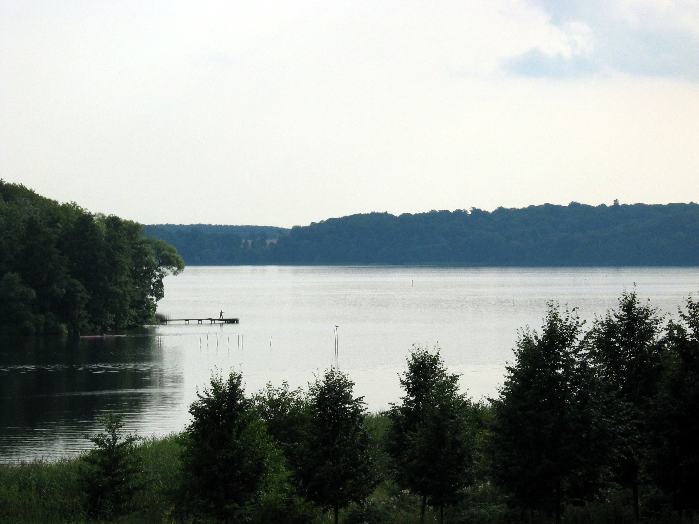 A wide, calm lake surrounded by dense green forests under a bright, overcast sky.