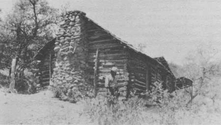 A black and white photograph of a historic, rustic log cabin with a stone chimney, situated in a dry, wooded area.