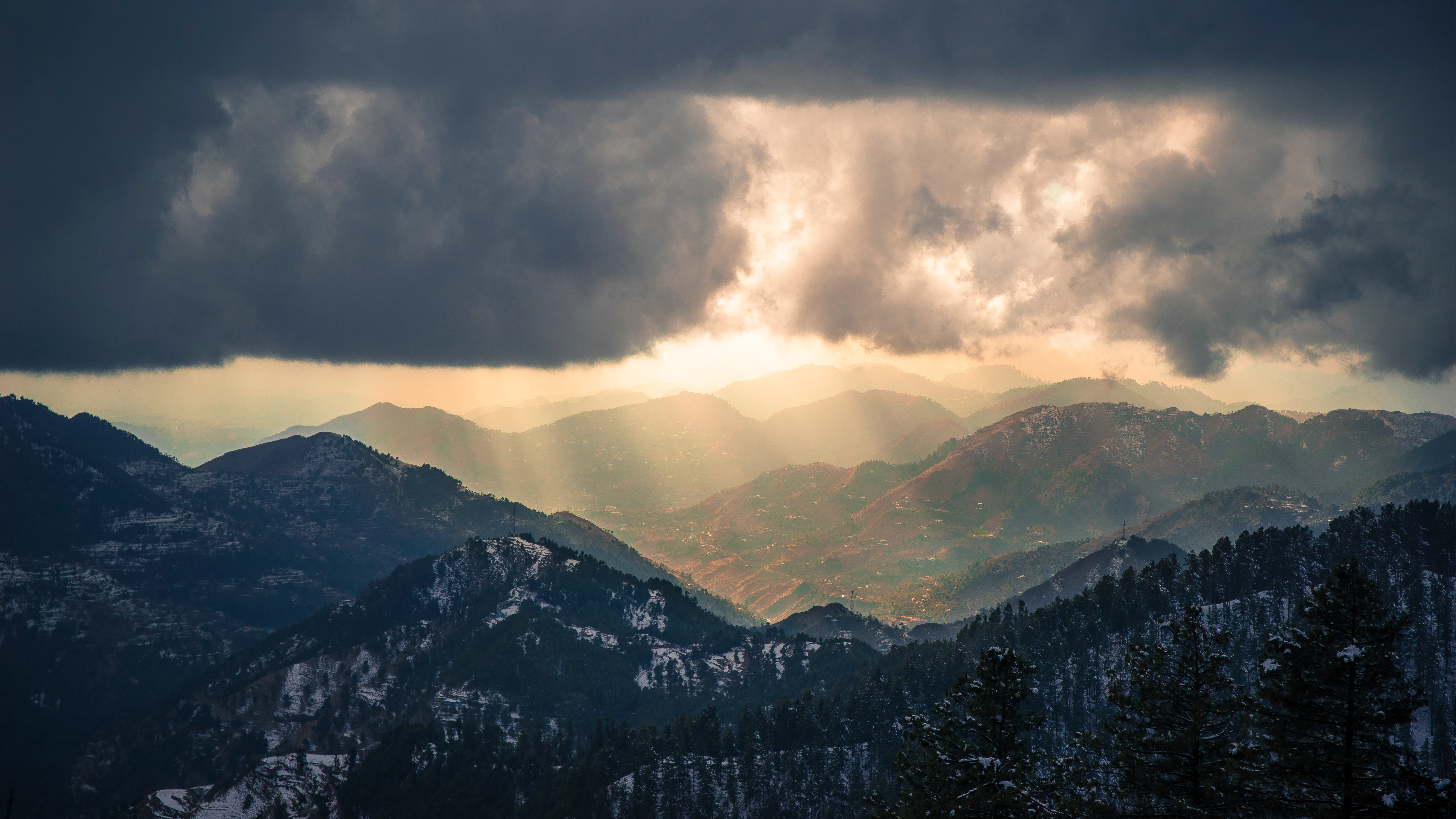 Una vista amplia y dramática del paisaje con cadenas montañosas en capas bajo un cielo pesado, oscuro y tormentoso, con rayos de sol que atraviesan las nubes para iluminar los valles.
