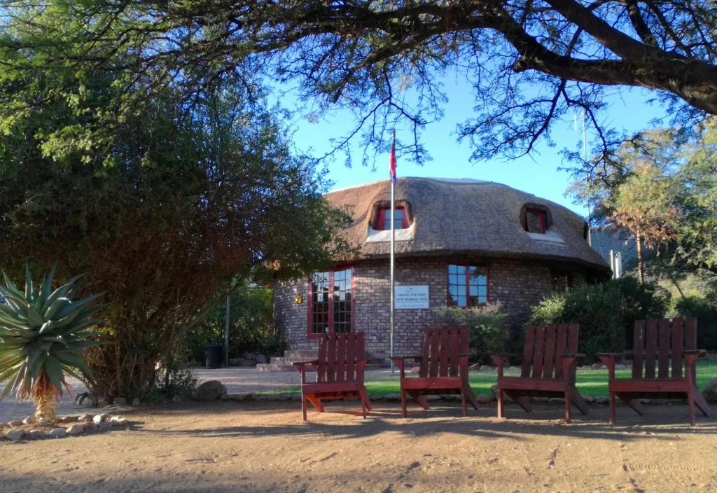 A round stone visitor center building with a thatched roof, fronted by a line of four wooden chairs under the shade of trees.