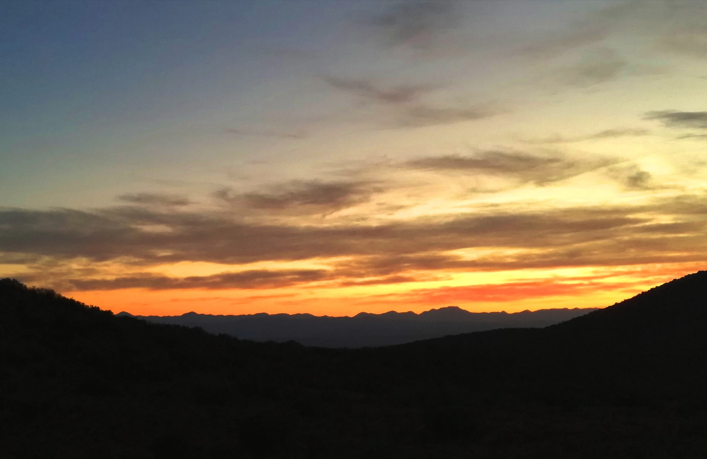 A vibrant sunrise with orange and yellow hues illuminating the sky above the dark silhouette of rolling mountain ridges.