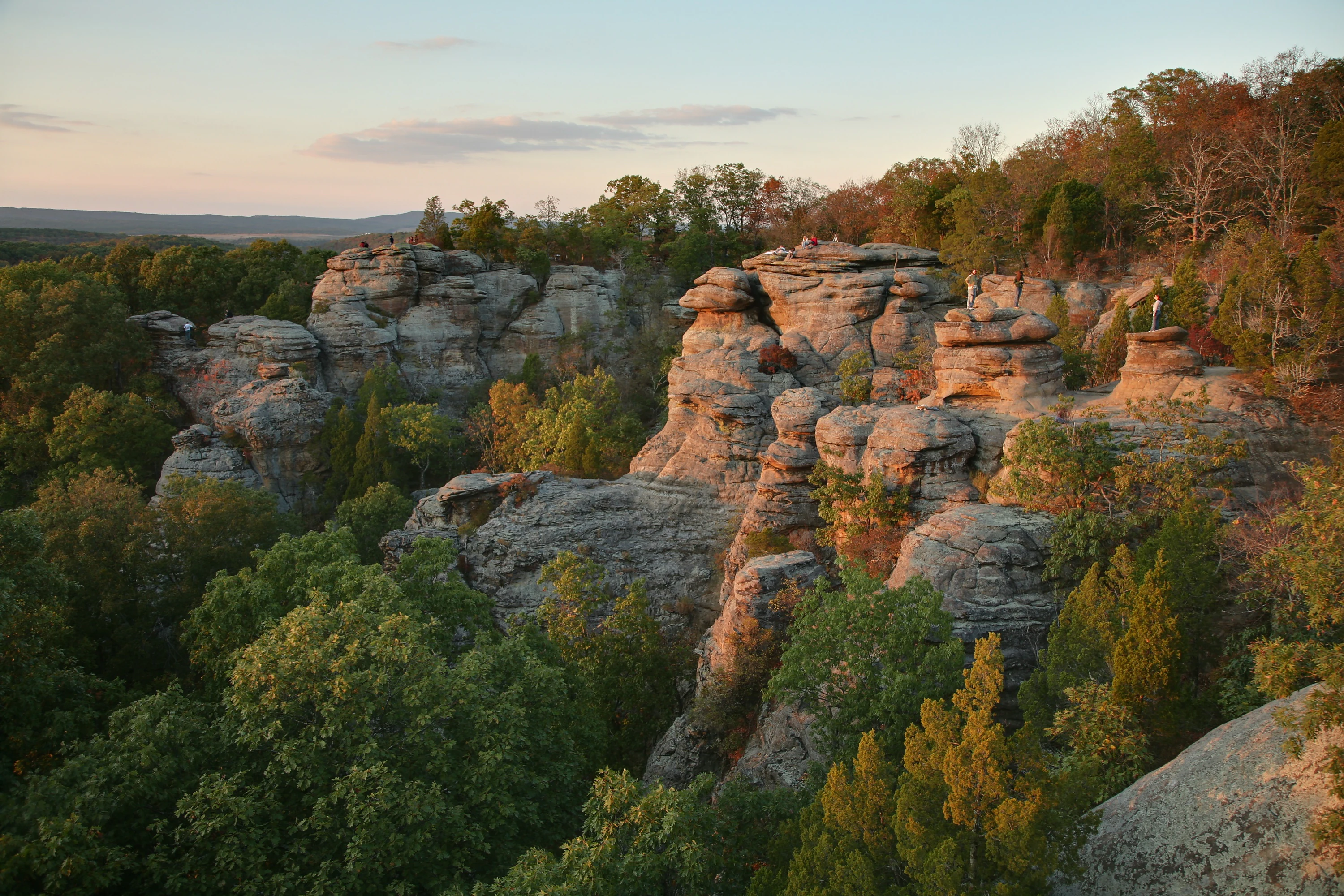 A wide angle landscape view of Garden of the Gods in Illinois at sunset, featuring large, weathered sandstone cliffs and rock outcrops surrounded by lush green and autumn-tinged deciduous trees.