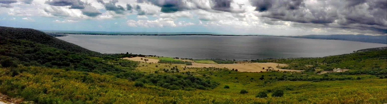 Panoramic landscape of a large lake surrounded by green hills under a partly cloudy sky.