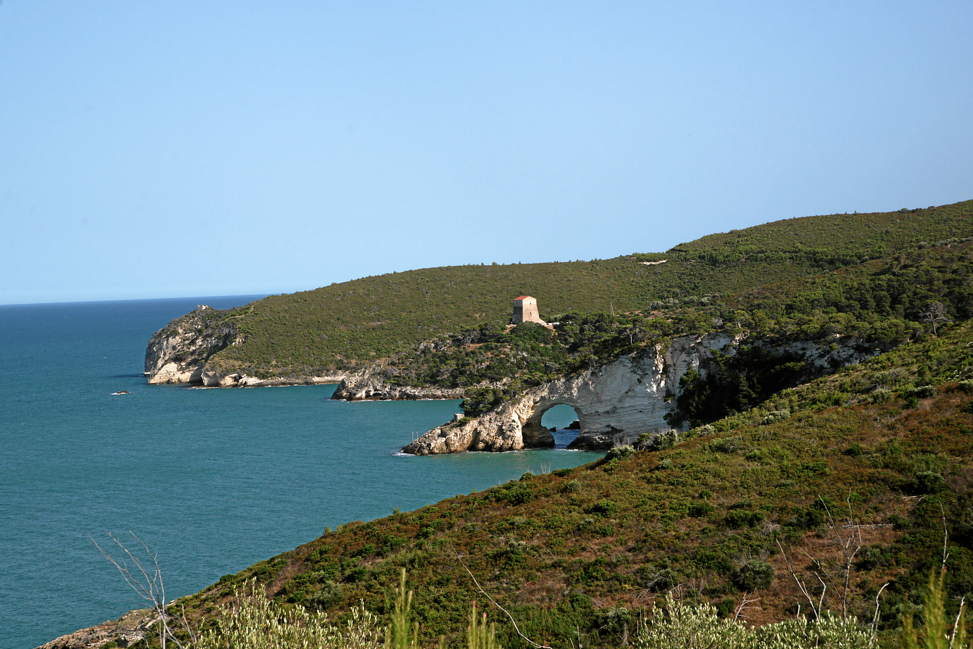 Coastal landscape featuring turquoise sea, rocky cliffs, a natural arch, and a small tower on a hillside