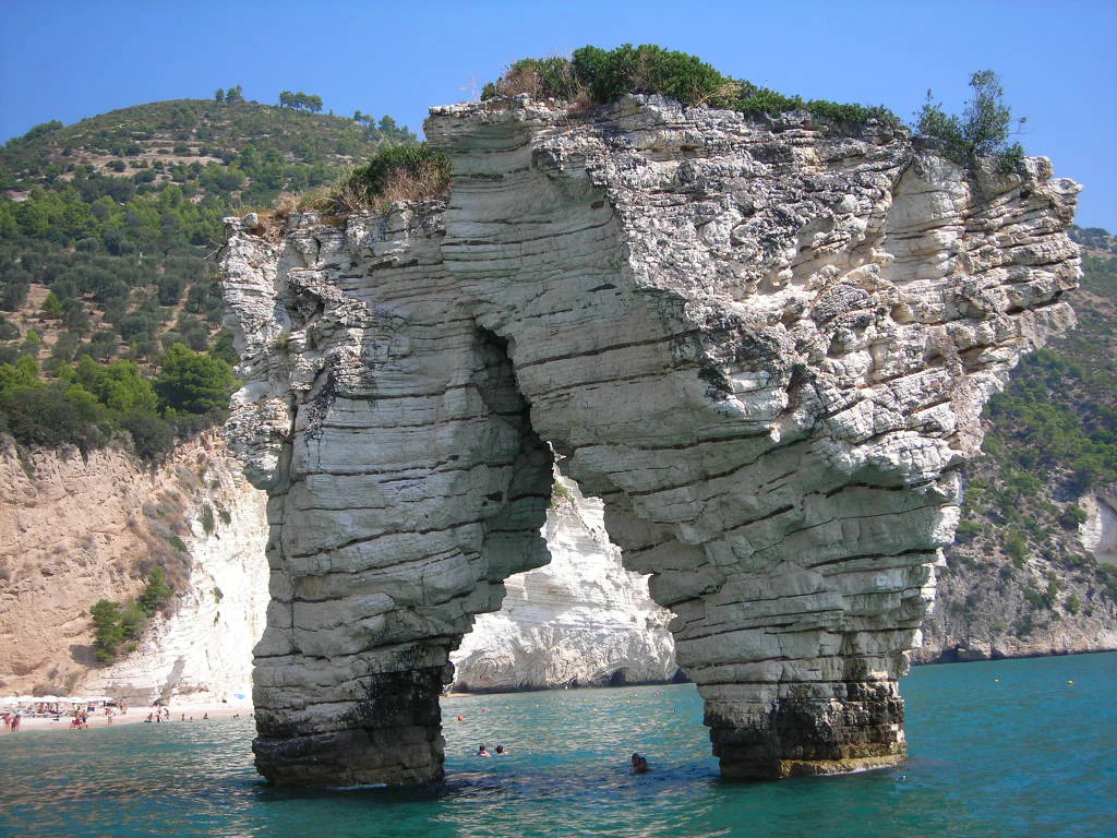 Large limestone sea stack with natural arch formation in turquoise water, beach with people in background, green hills on the horizon