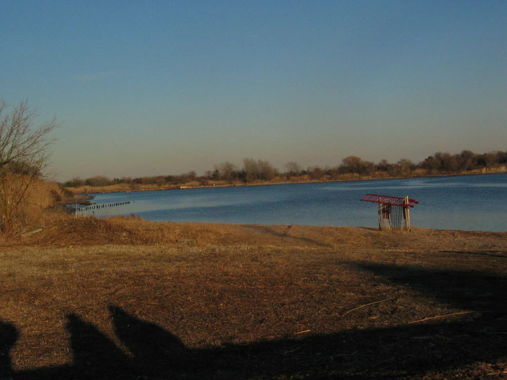 A wide, eye-level view of a calm bay with a brown, grassy shoreline in the foreground and a small, simple wooden shelter standing near the water's edge.
