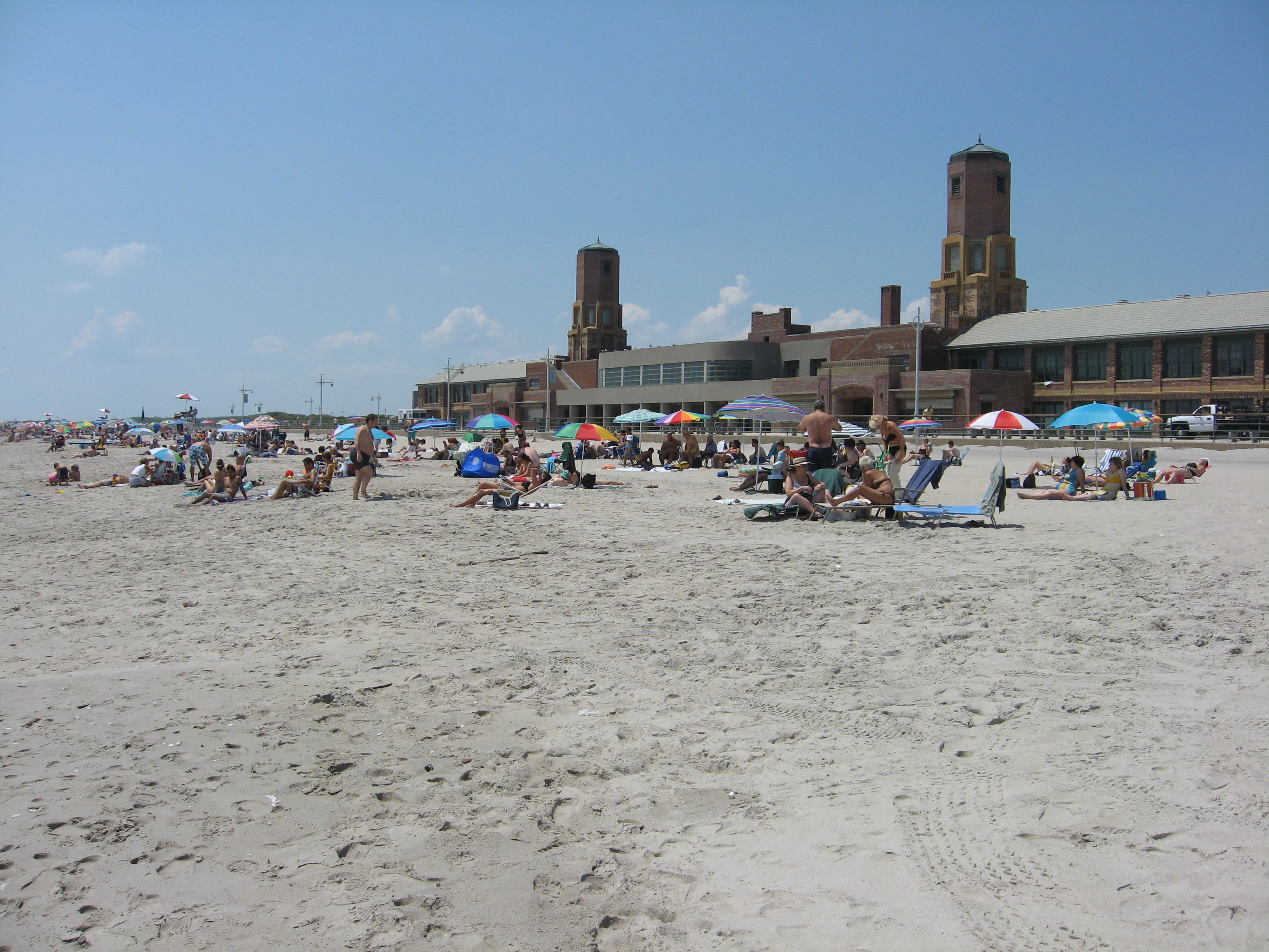 Wide view of a sandy beach with people lounging under colorful umbrellas, featuring a large brick historic bathhouse building with two towers in the background.