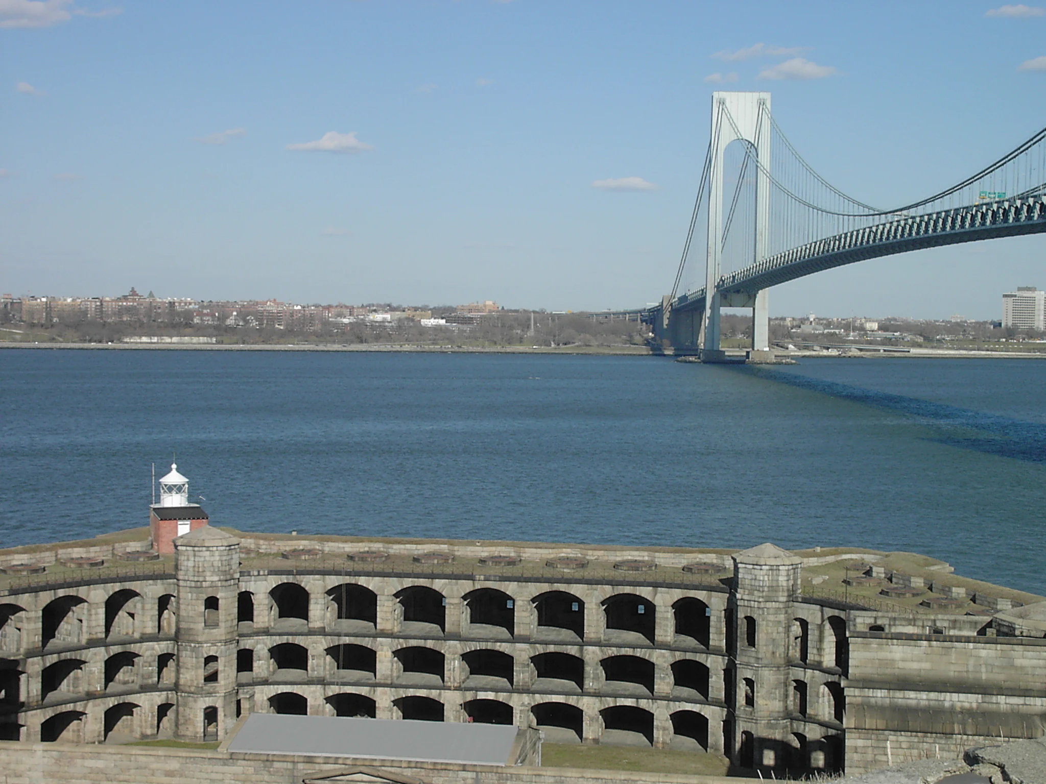A wide shot of the semicircular masonry walls of Fort Wadsworth in the foreground, with the massive Verrazano-Narrows Bridge spanning a large body of water in the background.