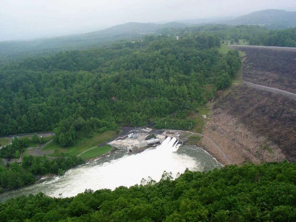 An aerial perspective shows a large rock-fill dam with a spillway discharging a powerful rush of white water into the river below, surrounded by dense green forest in the Appalachian mountains.