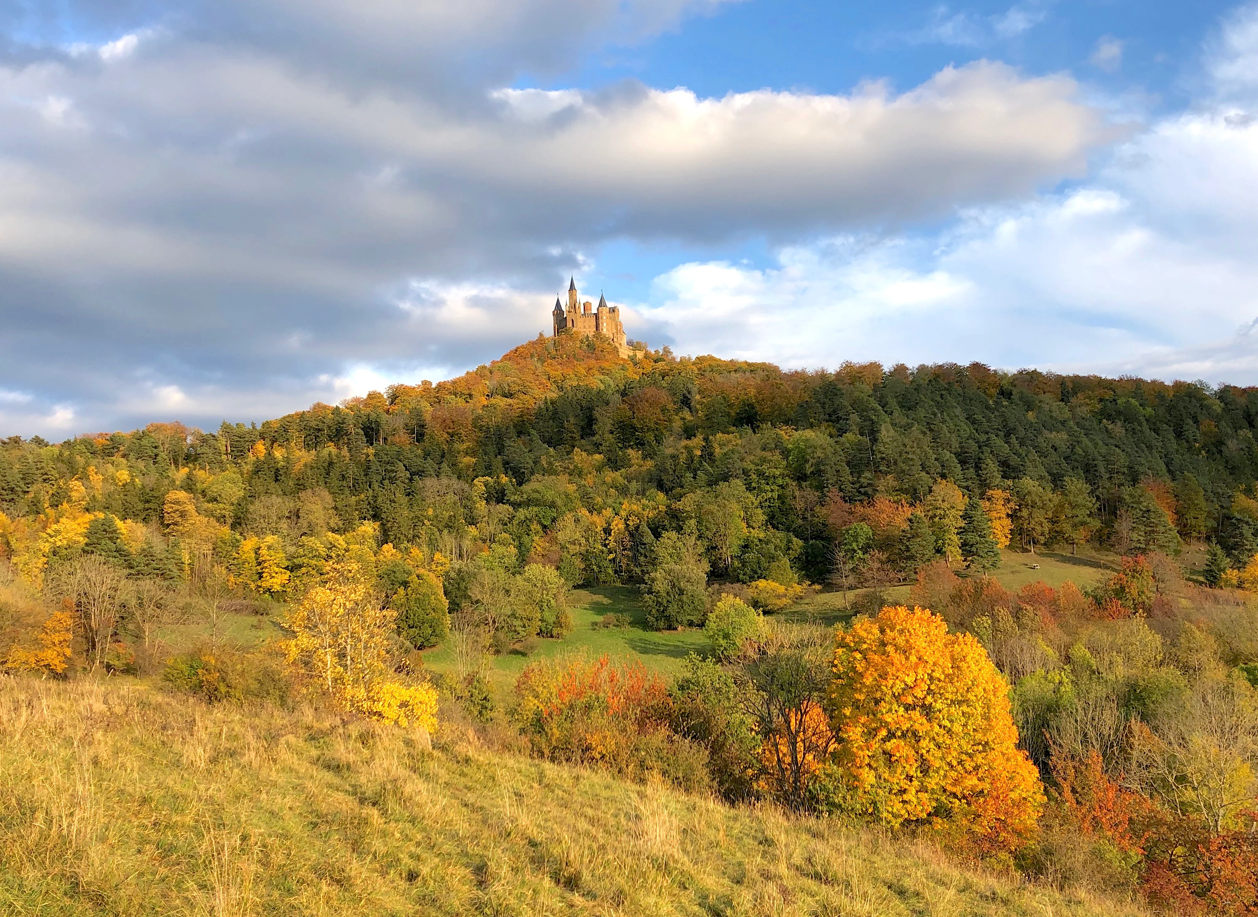 A wide view of a forested hillside with autumn foliage leading up to a stone castle on the hilltop under a partially cloudy blue sky.