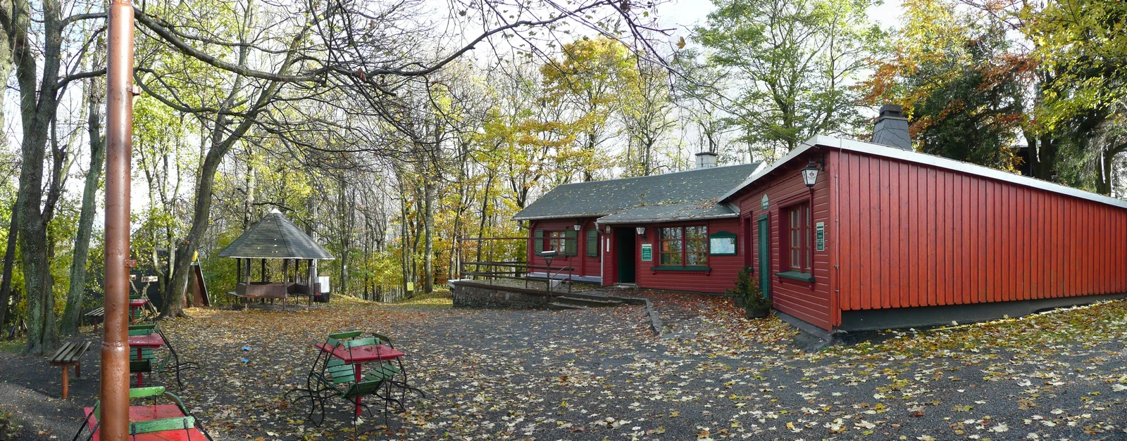 A red wooden mountain hut with a metal roof surrounded by autumn trees and fallen leaves on a paved summit area.