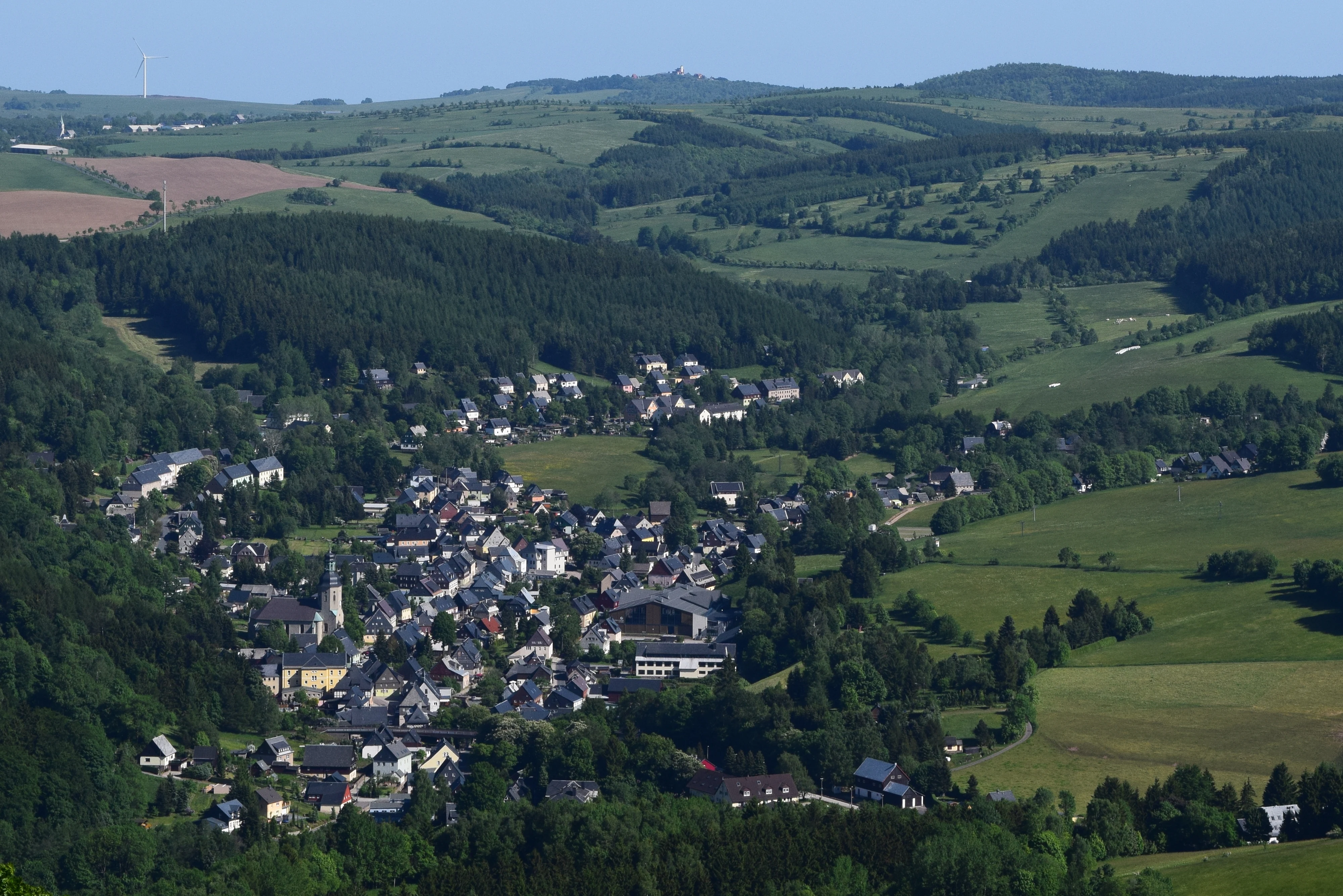 A wide, elevated view looking down into a green valley containing the small town of Geising, surrounded by rolling hills and patches of forest.
