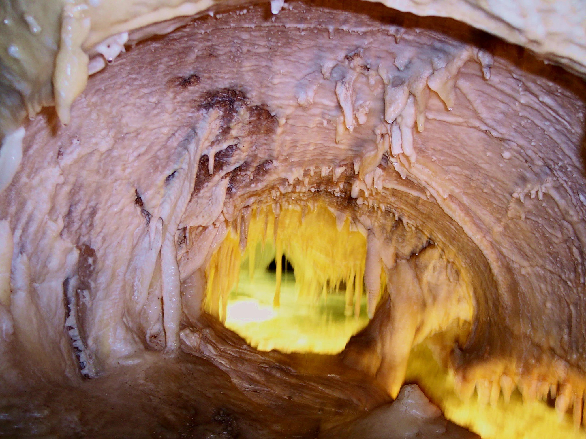 Vista a baja altura dentro de una cueva con paredes rocosas texturizadas y estalactitas, que muestra un pequeño lago subterráneo brillantemente iluminado en el centro.