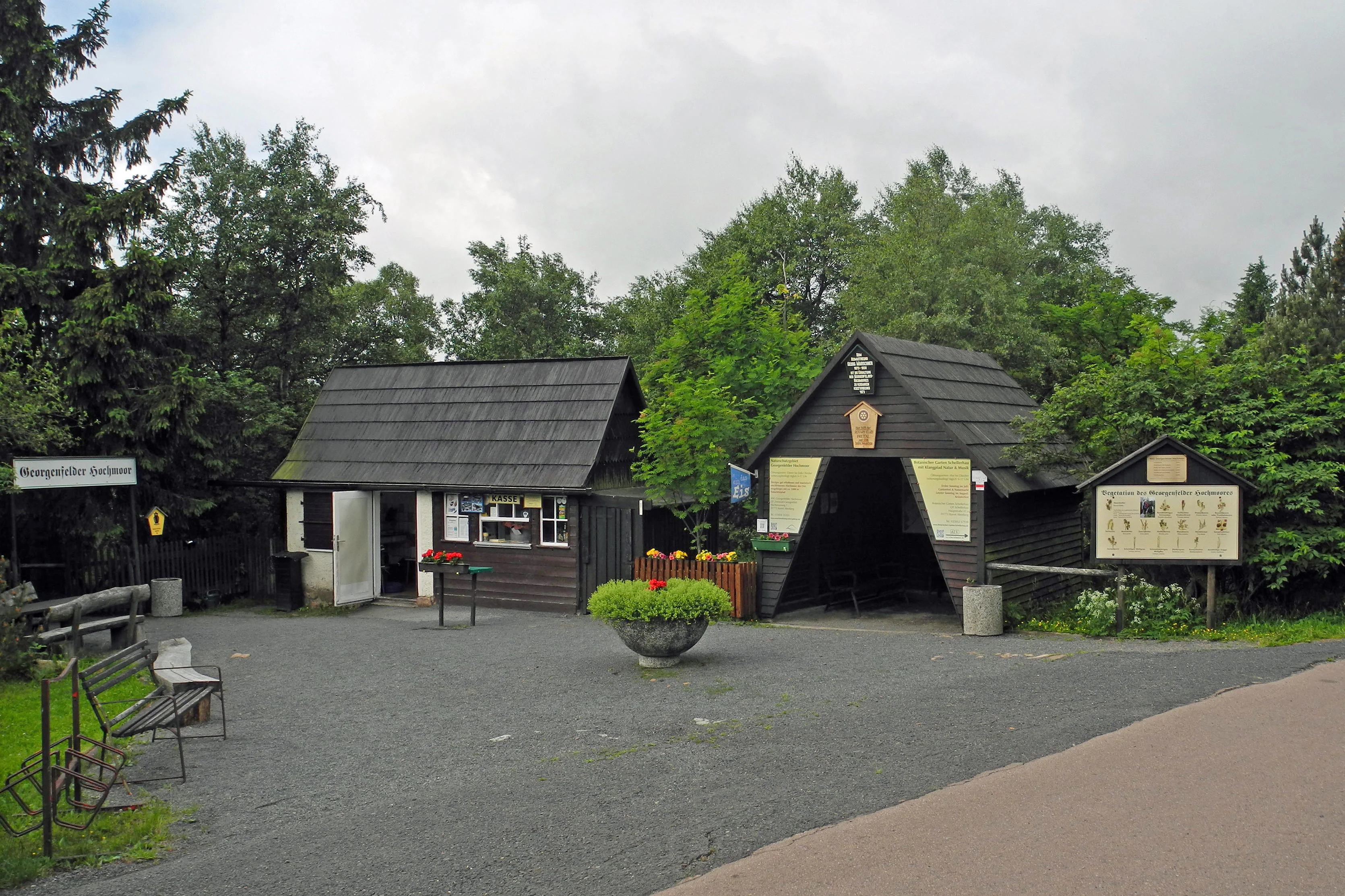 A gravel-covered entrance area with two wooden buildings and information signs marking the entrance to the Georgenfelder Hochmoor, surrounded by dense trees.