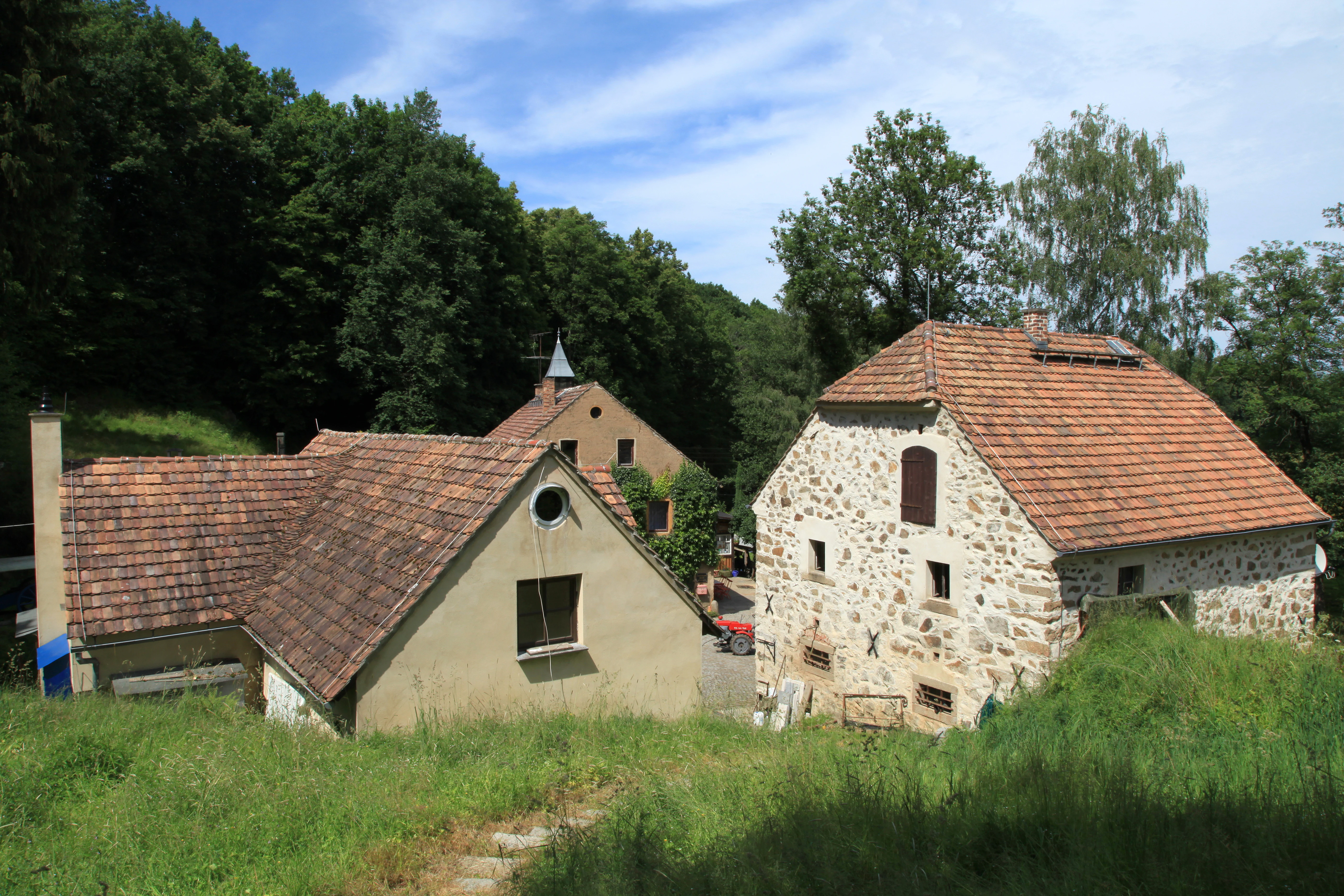 A group of rustic stone and masonry buildings with red tile roofs situated in a green, wooded valley area.