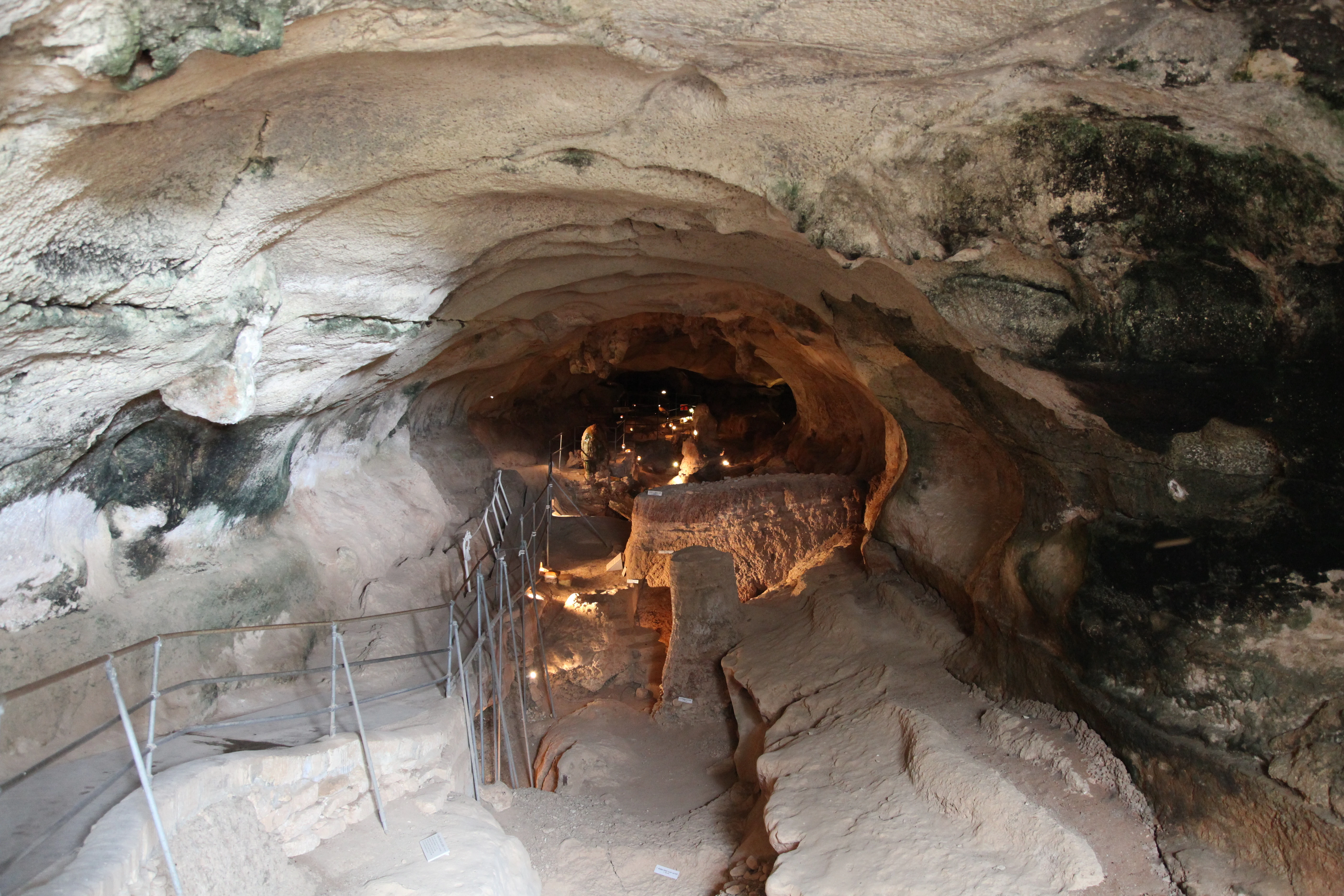 A wide angle view looking into a limestone cave with a lit walking path, metal railings, and natural rock walls.