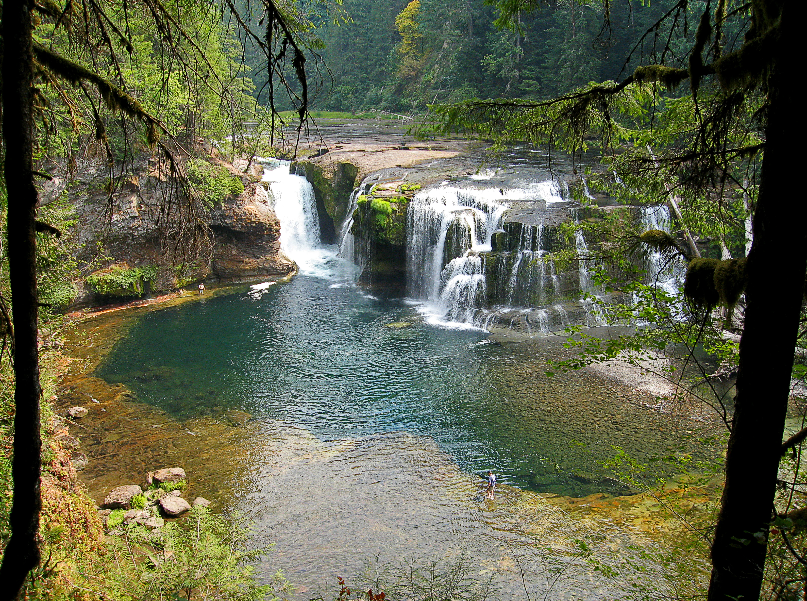 A wide, multi-tiered waterfall cascading over dark basalt rock into a calm, turquoise plunge pool, framed by lush overhanging forest foliage.