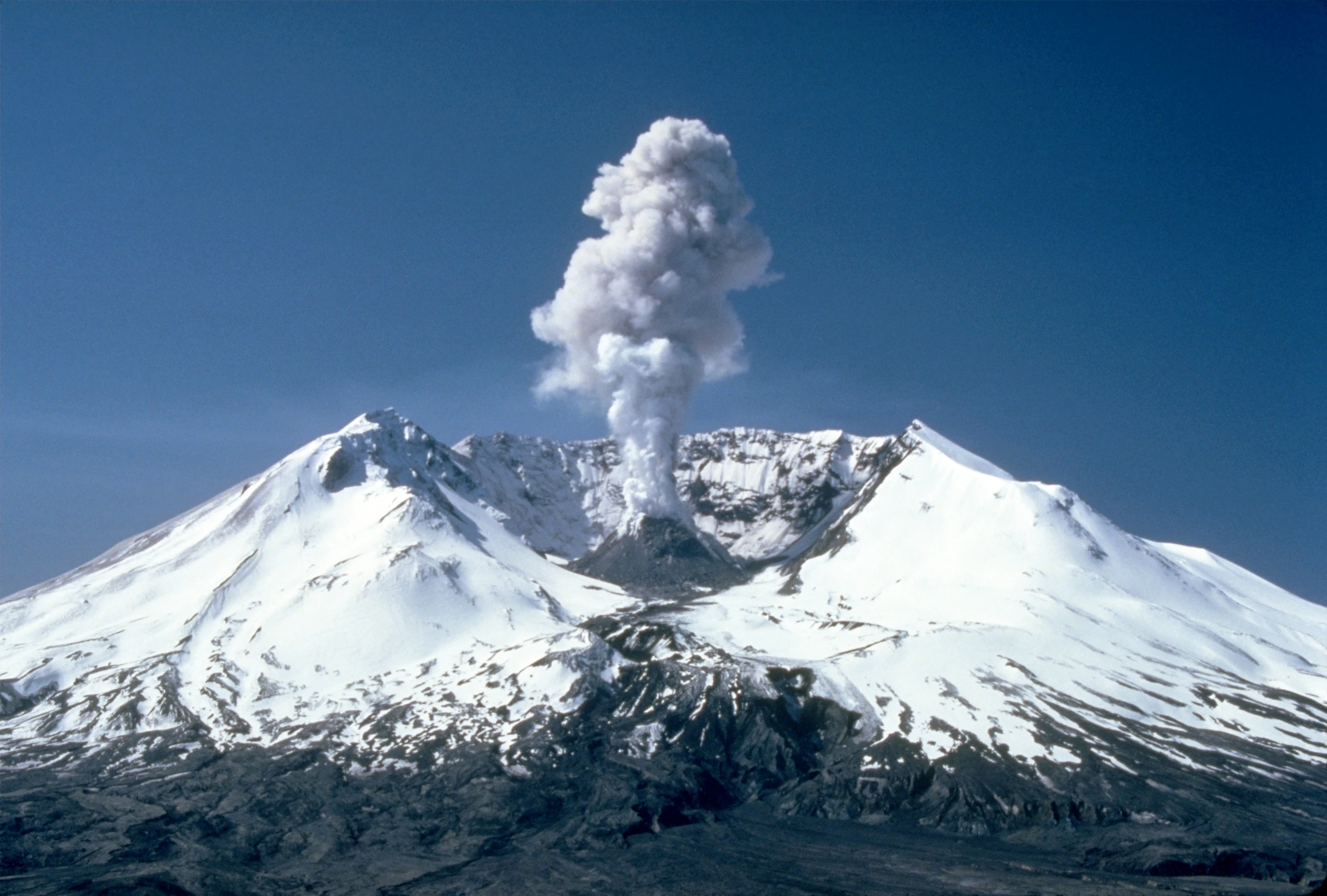 A view of the snow-covered peak of Mount St. Helens with a large plume of steam and ash rising from the crater against a clear blue sky.