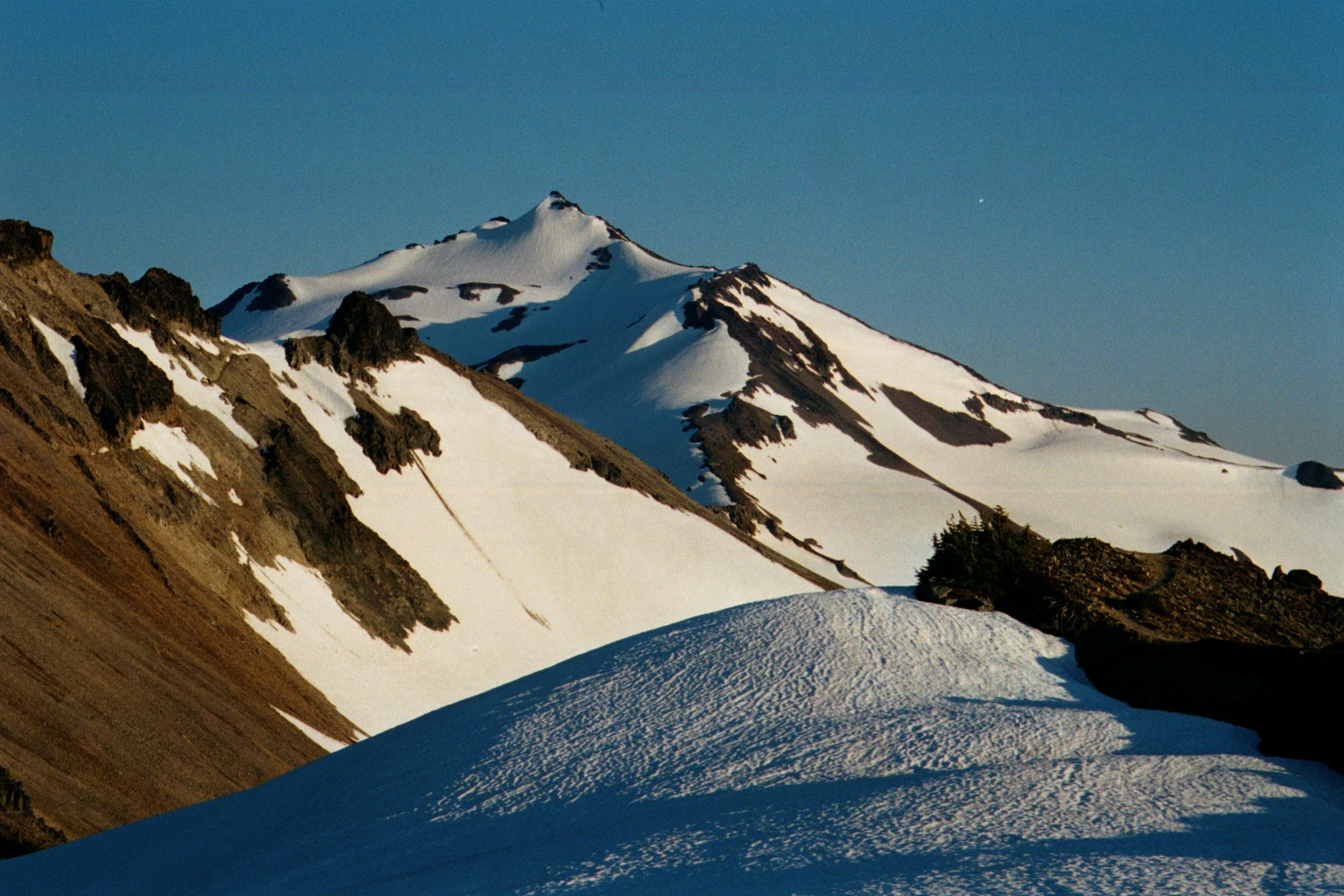 A snowy, craggy mountain peak rises against a clear blue sky, with a foreground covered in snow drifts and rocky slopes.