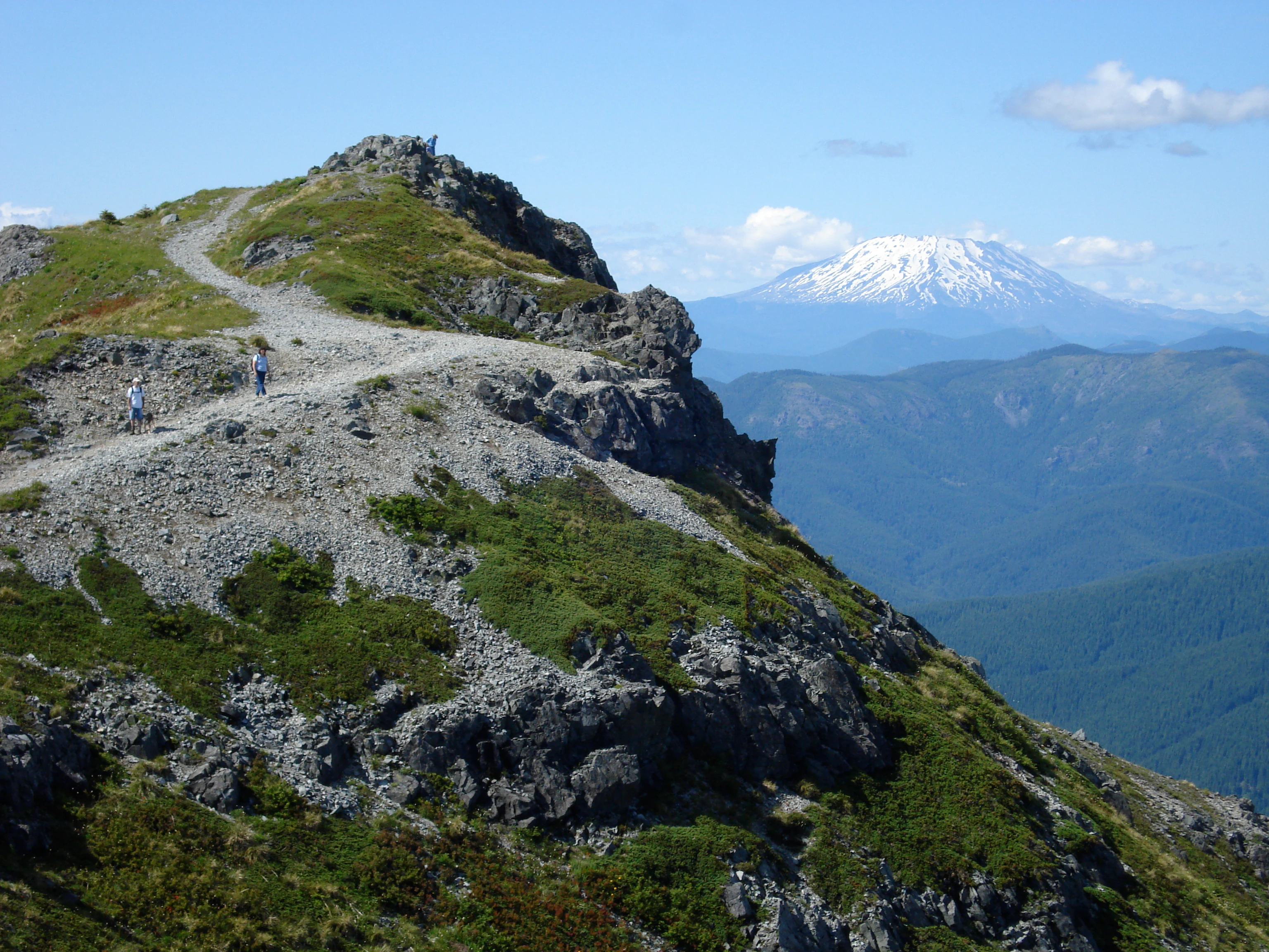 A dirt hiking trail follows a rocky ridgeline toward a mountain summit, with two hikers walking along the path and a large, snow-covered volcanic peak visible in the background under a blue sky.