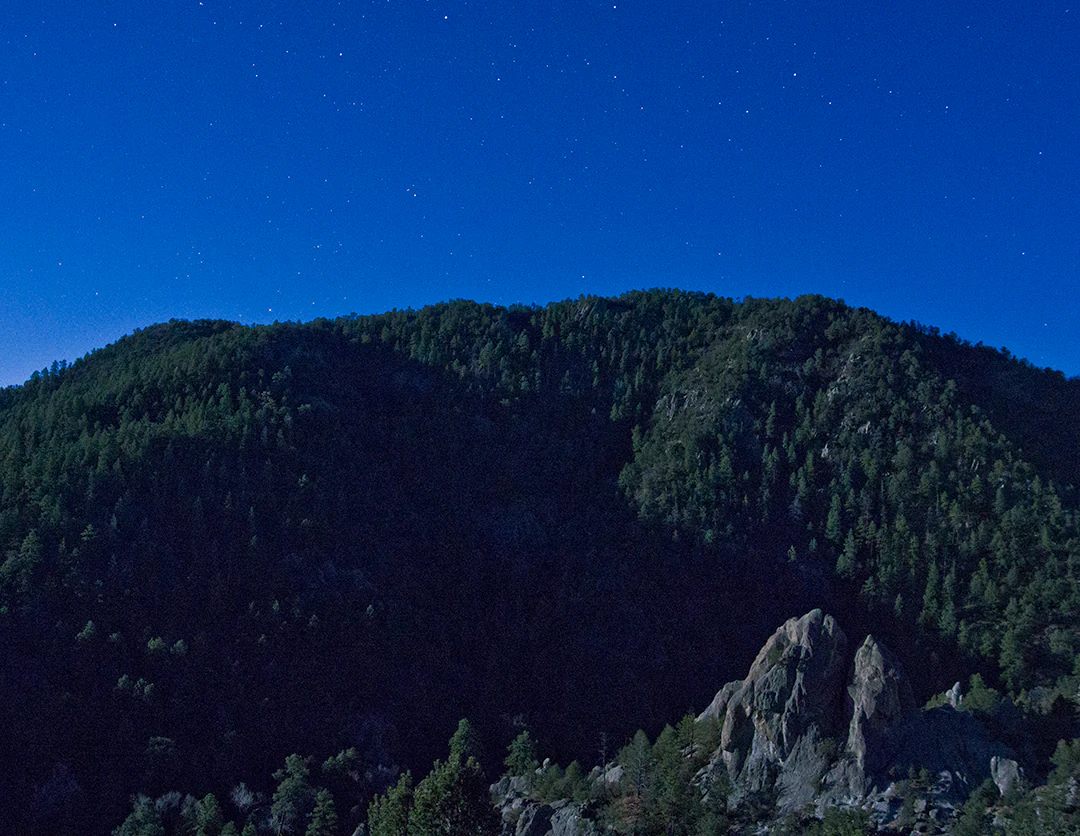 A moonlit landscape showing a dark, forested mountain range against a clear night sky with visible stars.